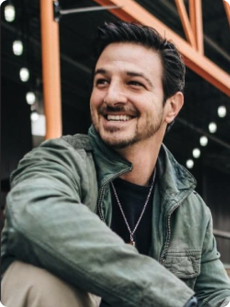 A smiling man with dark hair and a beard, wearing a green jacket and a black shirt, sitting outdoors under a structure with orange beams and string lights.