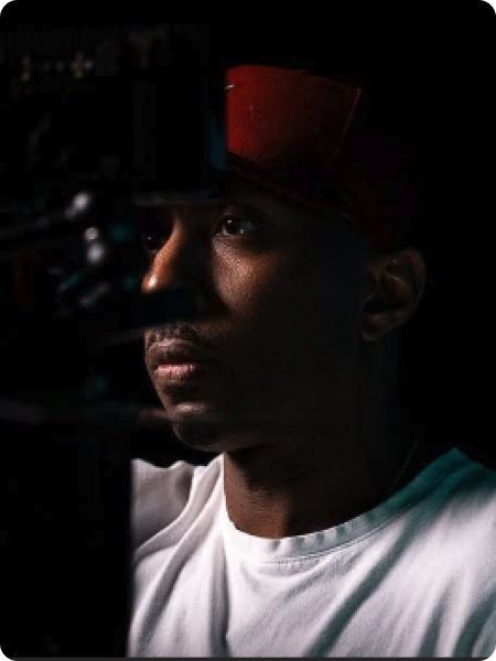 A man wearing a red cap and a white shirt, looking to the side with a serious expression, in a dark setting with minimal lighting.