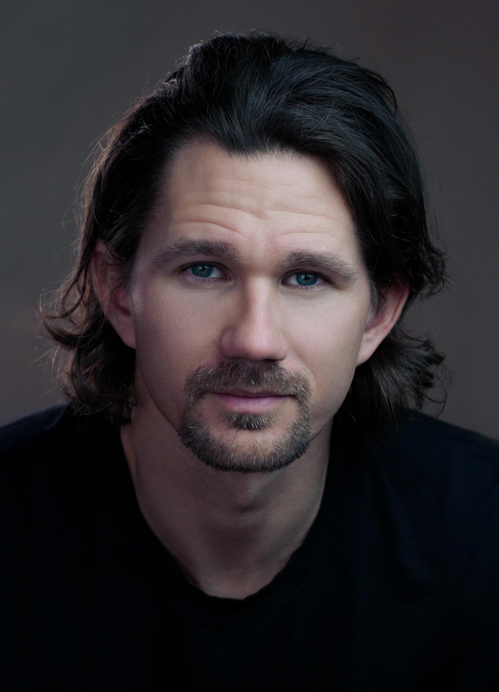 Close-up portrait of a man with long dark wavy hair, blue eyes, and a goatee, wearing a black shirt against a dark background.