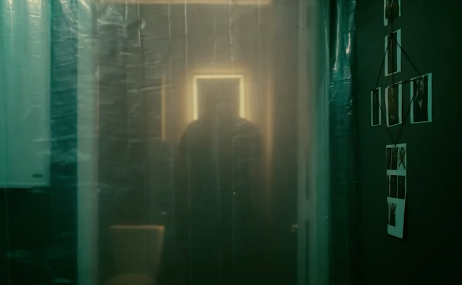 Reflection of a person taking a photo in a semi-transparent curtain or plastic sheet with a neon square light in the background and a wall decorated with photos on the right side.