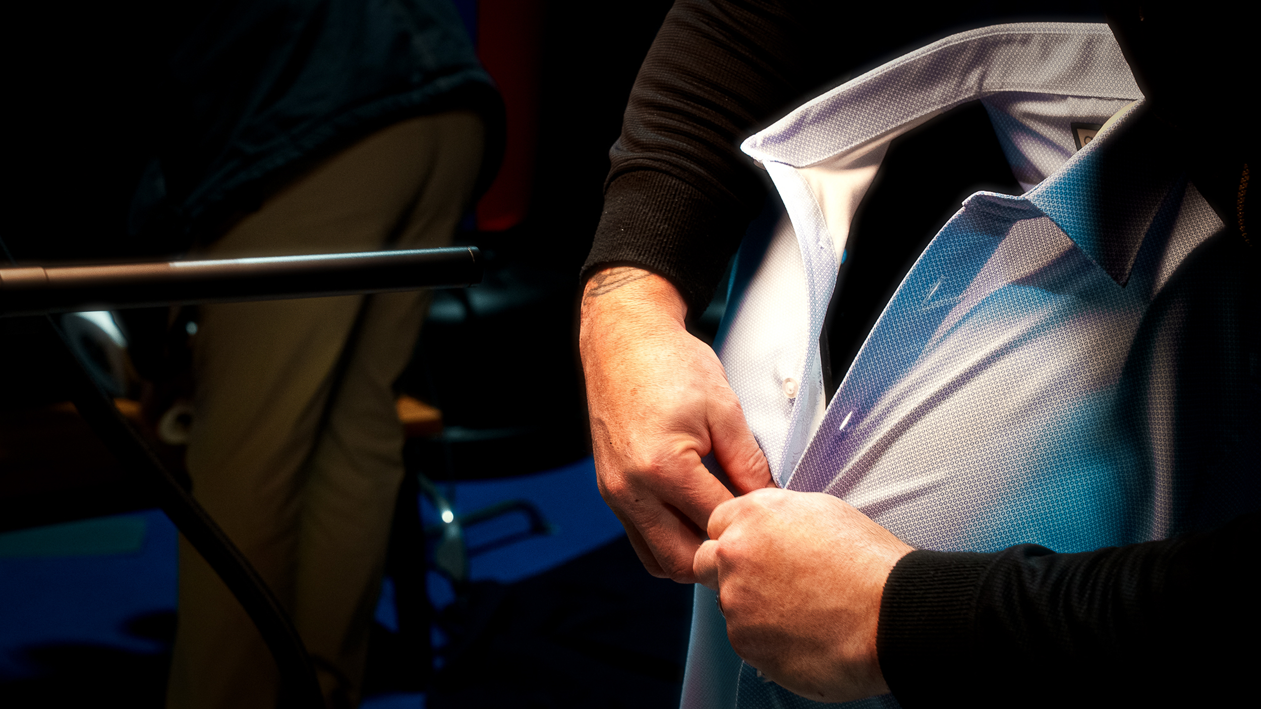 A person is adjusting their dress shirt cuffs under a black sweater, while another person in the background is leaning over a table.