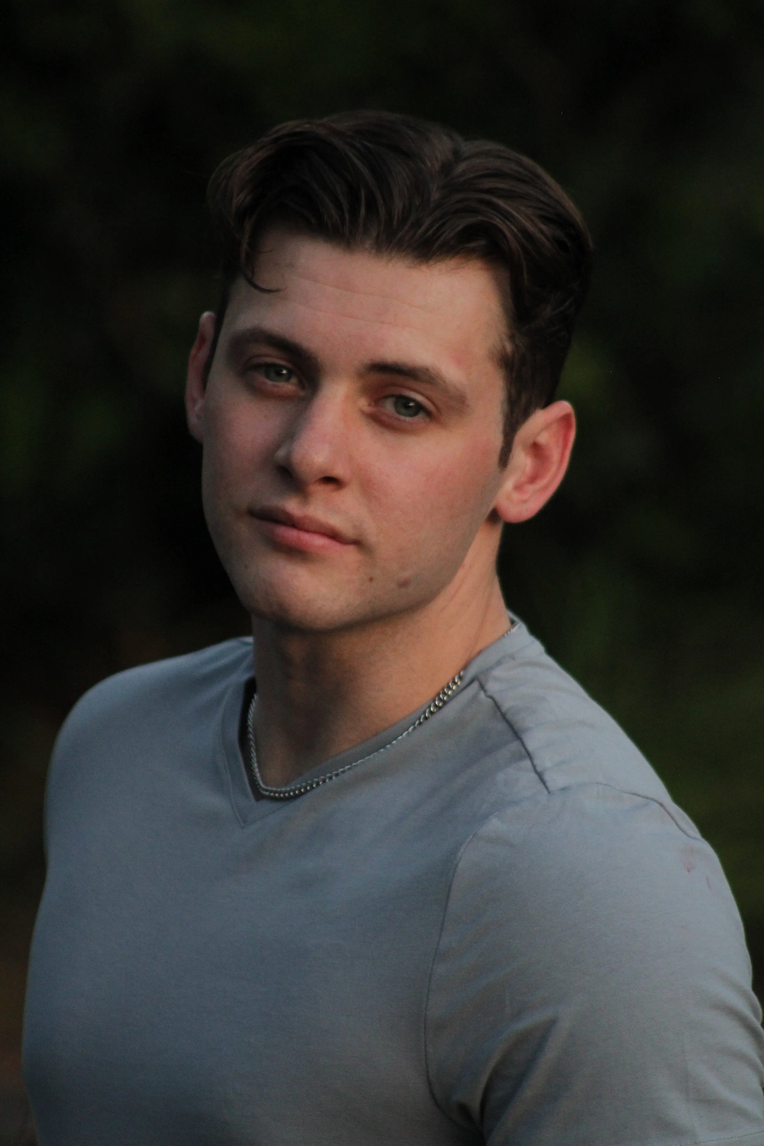 A young man with dark hair, wearing a gray shirt and a chain necklace, posing outdoors with a dark, blurred background.