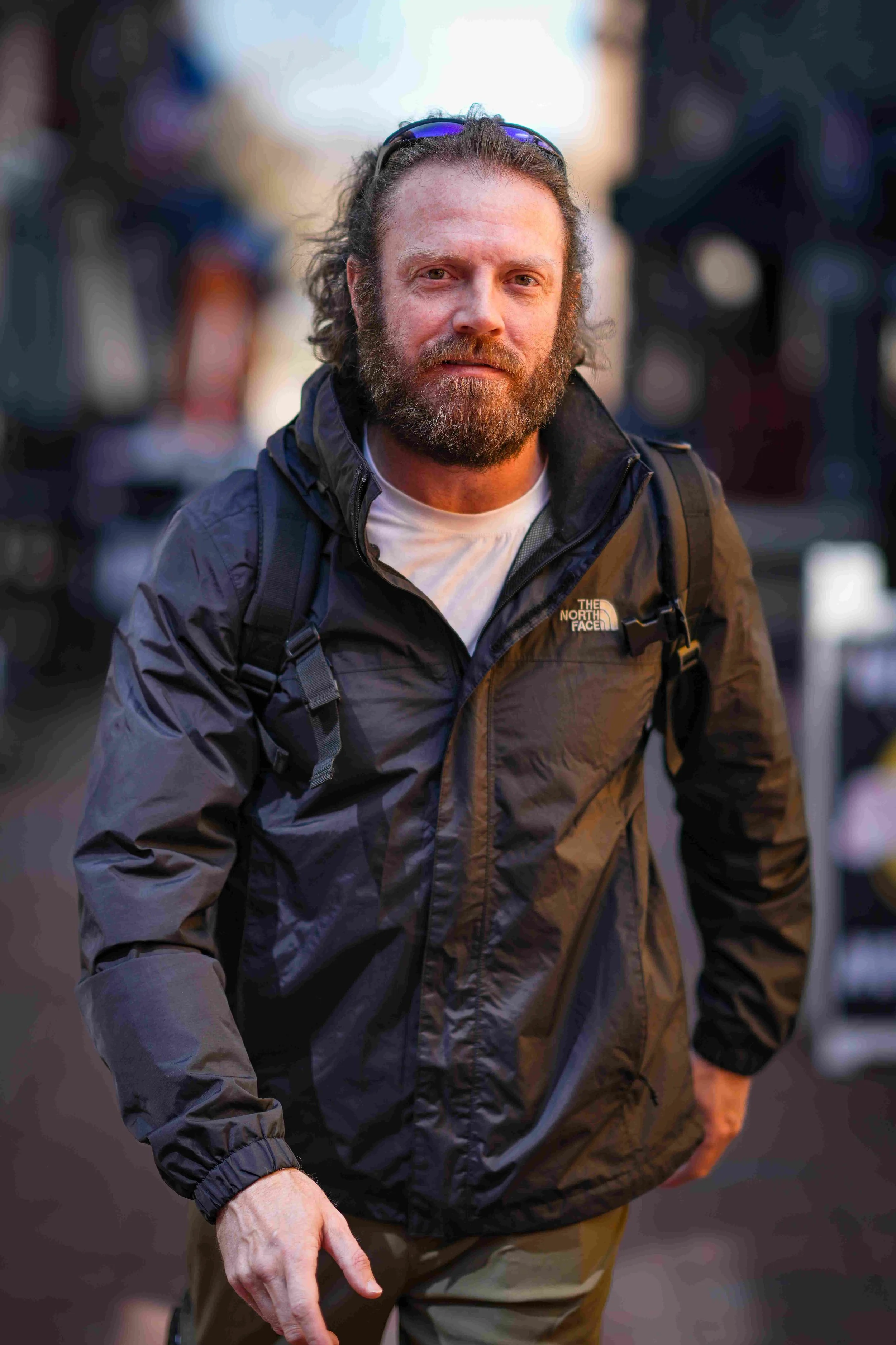A bearded man wearing a black North Face jacket, sunglasses on his head, and a white shirt underneath, standing outdoors during sunset or dusk with a blurred background.