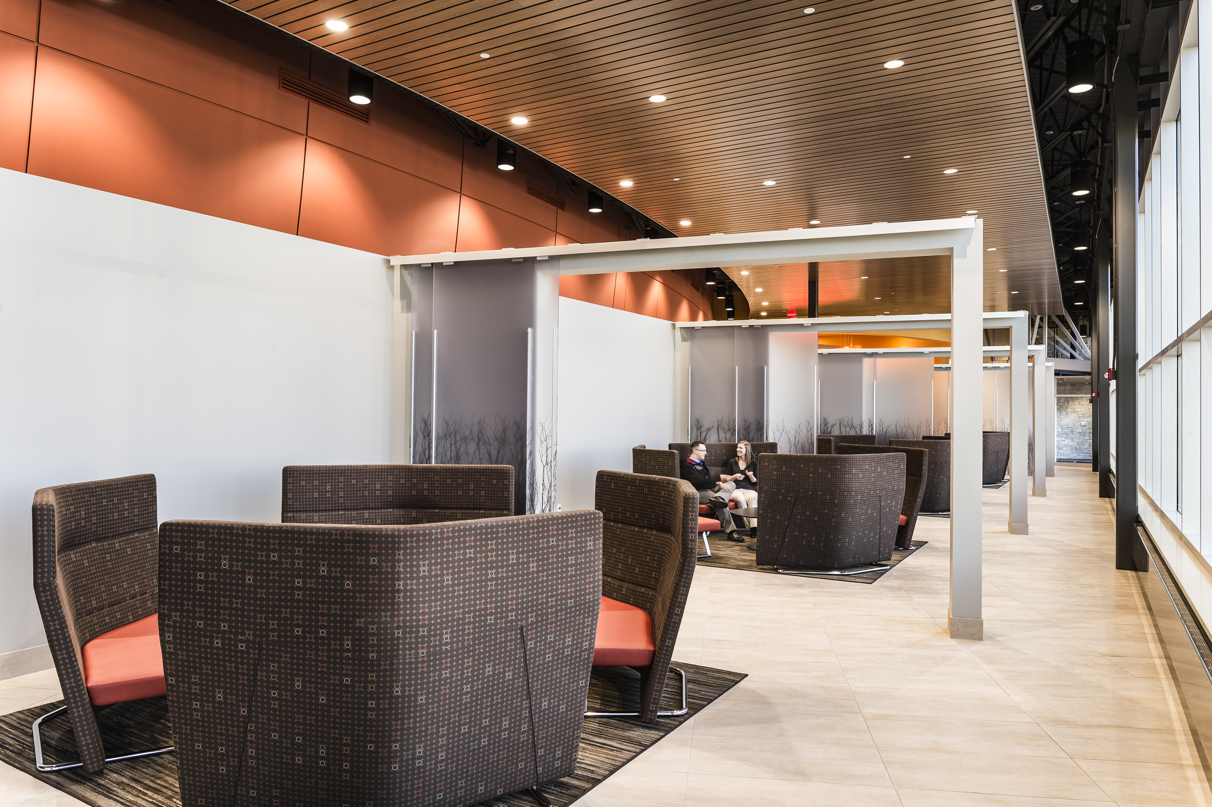 Modern interior of a lobby or waiting area with polished wood and tile flooring, large windows, curved red metal wall, and a yellow ceiling with circular red and white lighting fixtures. There are chairs, tables, and a person walking through the spac
