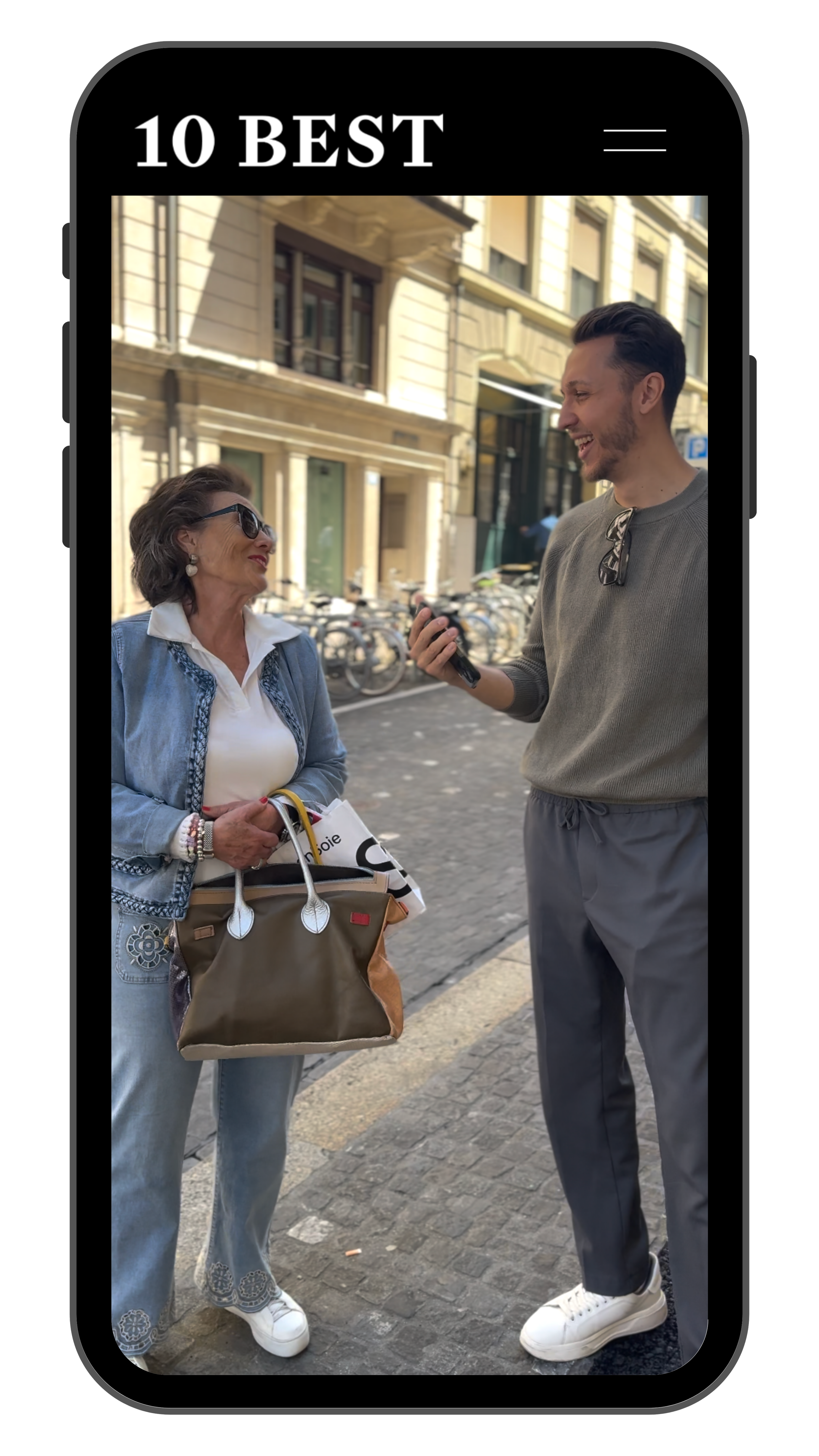 Zwei Menschen stehen auf der Straße, einer ist eine ältere Frau mit Sonnenbrille, einer ist ein junger Mann, der aussieht, als ob er mit dem Smartphone spricht, im Hintergrund Fahrräder und historische Gebäude.