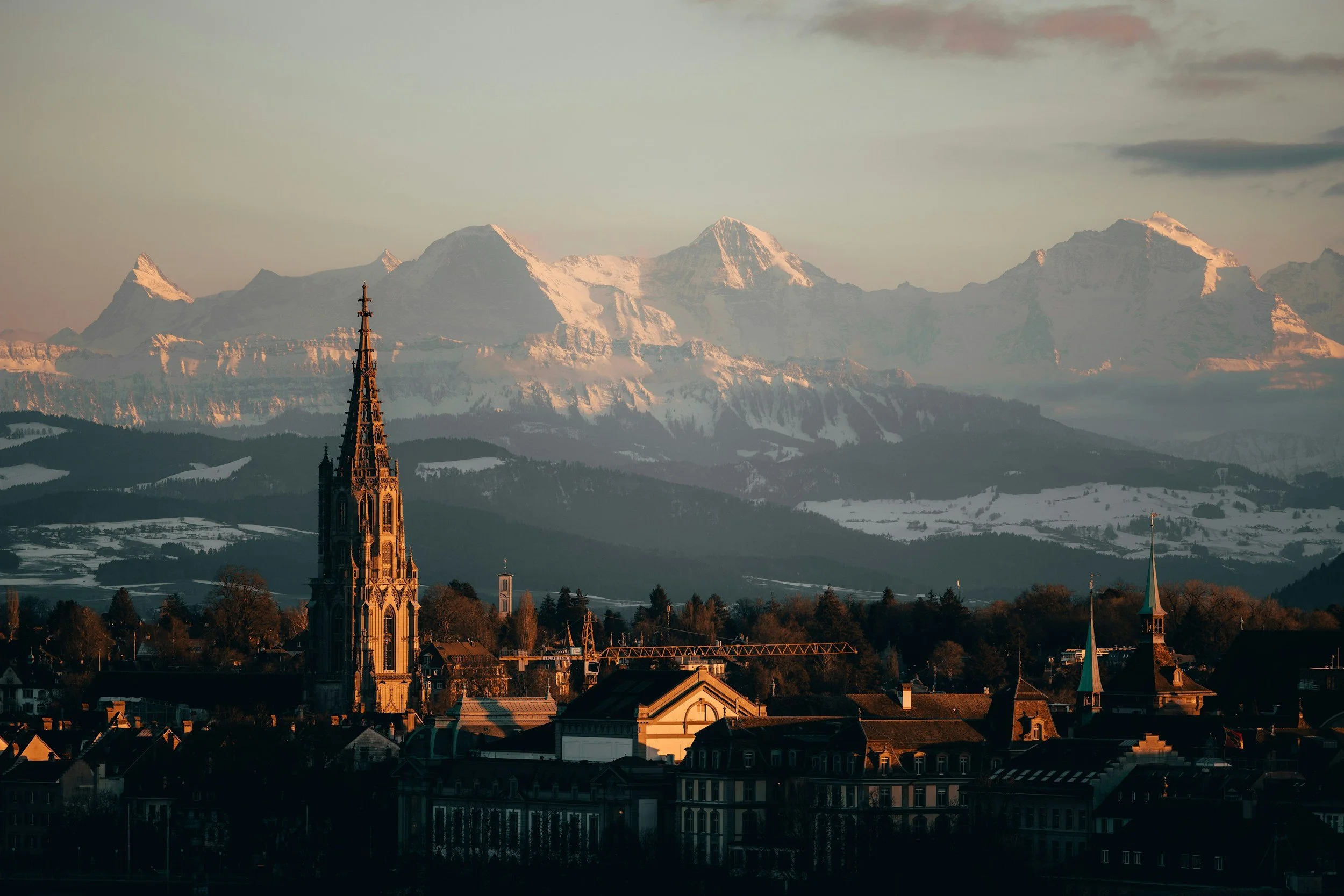 Blick auf eine Stadt mit gotischer Kirche im Vordergrund und verschneiten Bergen im Hintergrund bei Sonnenuntergang.