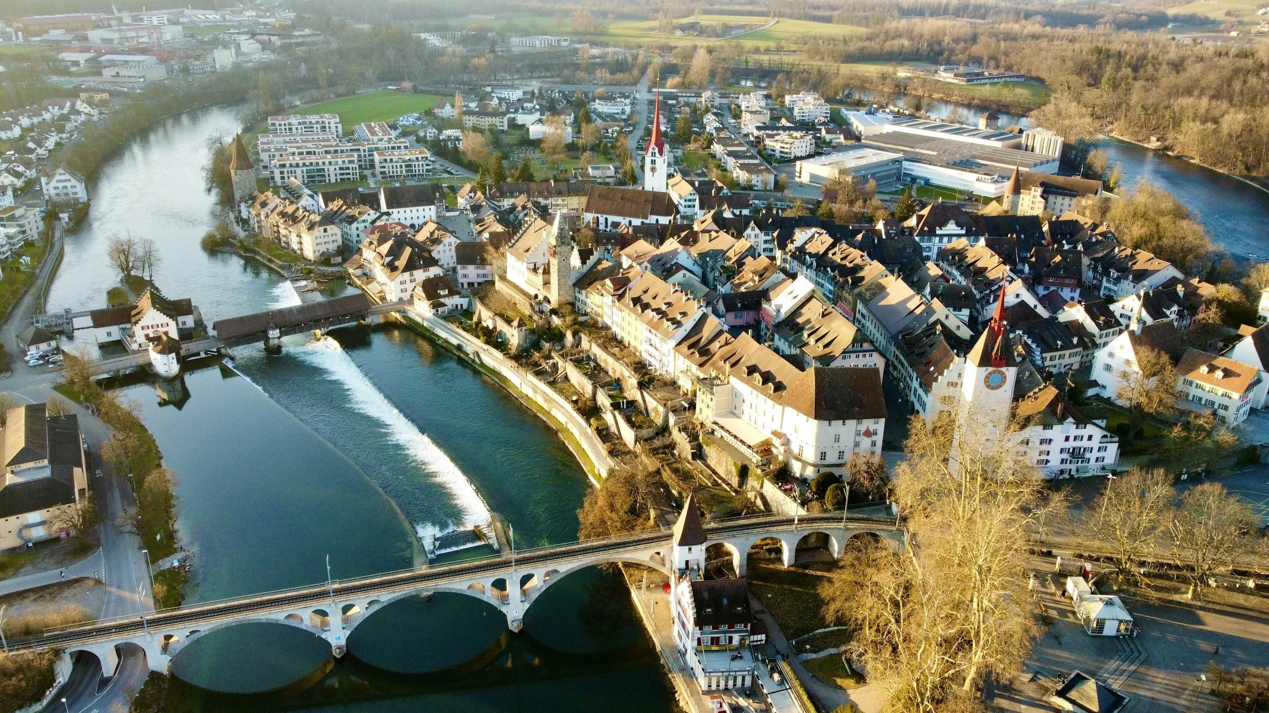 Luftaufnahme einer kleinen Stadt mit Fluss, Brücken, historischen Gebäuden und modernen Gebäuden, umgeben von Landschaft und Bäumen im Herbst.
