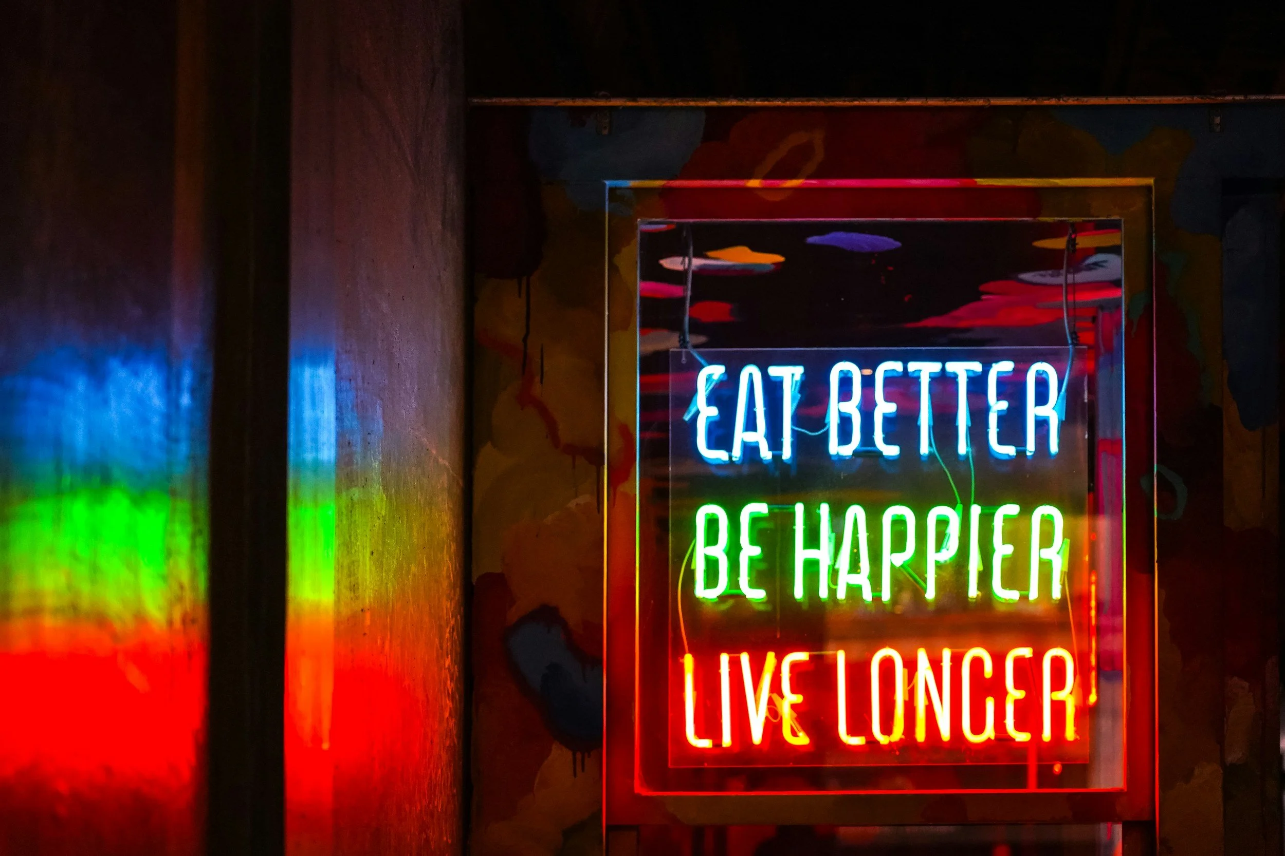 Neon-Wandschild mit den Worten 'Eat Better', 'Be Happier' und 'Live Longer' in Blau, Grün und Rot, mit reflektierenden Farben an der Wand.