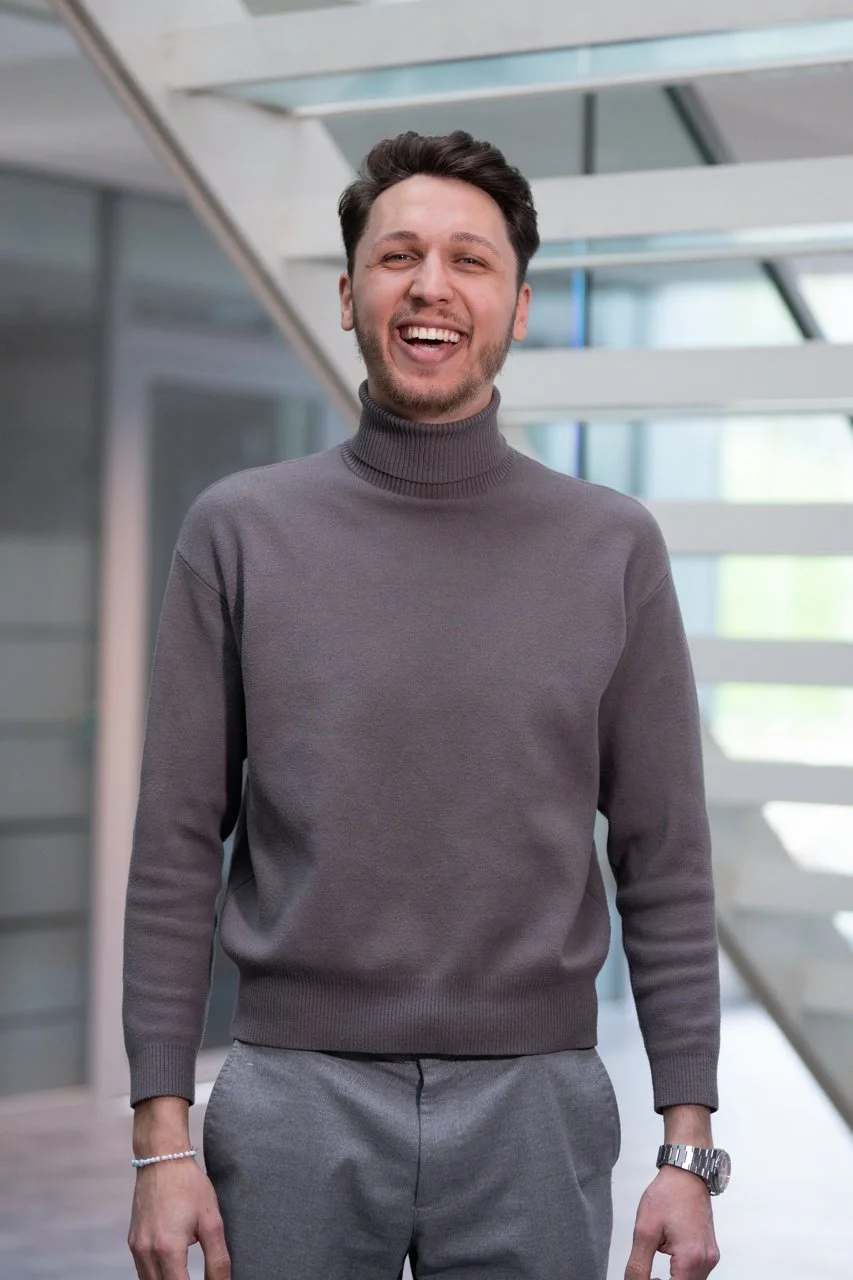Lächelnder Mann in grauem Rollkragenpullover und grauen Hosen, stehend in modernem Gebäude mit Glas und Stahl.