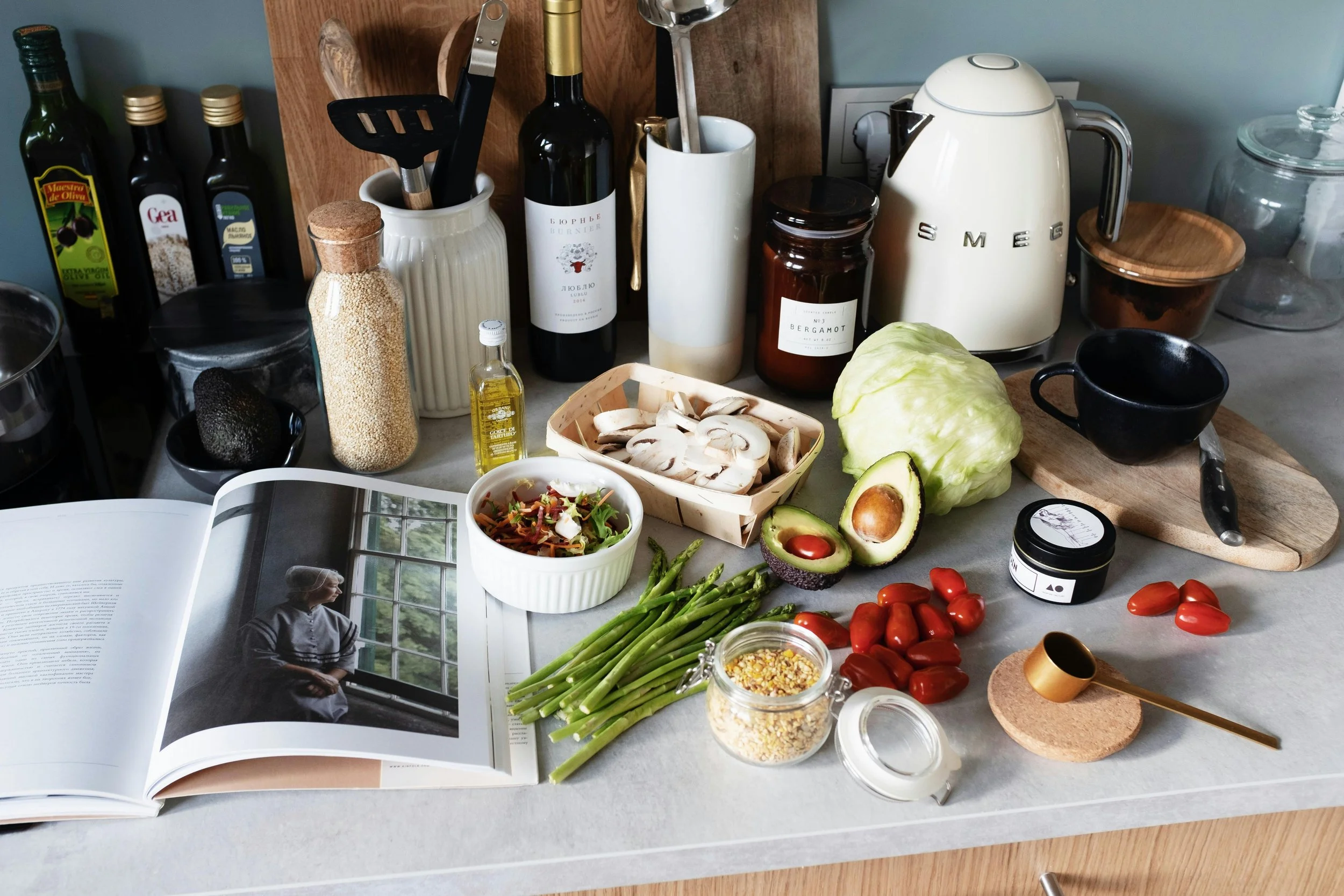 Häkelkorb mit geschnittenen Champignons, Avocado, Tomaten, grüne Spargelstangen, Salat in einer weißen Schüssel, offene Buchseite, Flasche mit Körnern, Ölflasche, dunkle Avocado, schwarzer Espresso im Becher, schwarze Küchenutensilien, Korken, Herd mit Küchenutensilien, elektrische Wasserkocher, Lebensmittelbehälter, Marmeladenglas, Schneidbrett mit Messer, weißer Kaffeekanne, Glas mit Körnern, offene Buchseite, Küchenregal im Hintergrund.