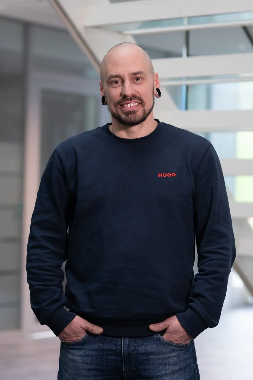 Junge Mann mit Glatze, Bart und großen Ohrringen, trägt einen schwarzen Pullover mit rotem Schriftzug 'HUGO', steht in modernem Gebäude mit Glas und Metall, lächelnd, Hände in den Taschen.