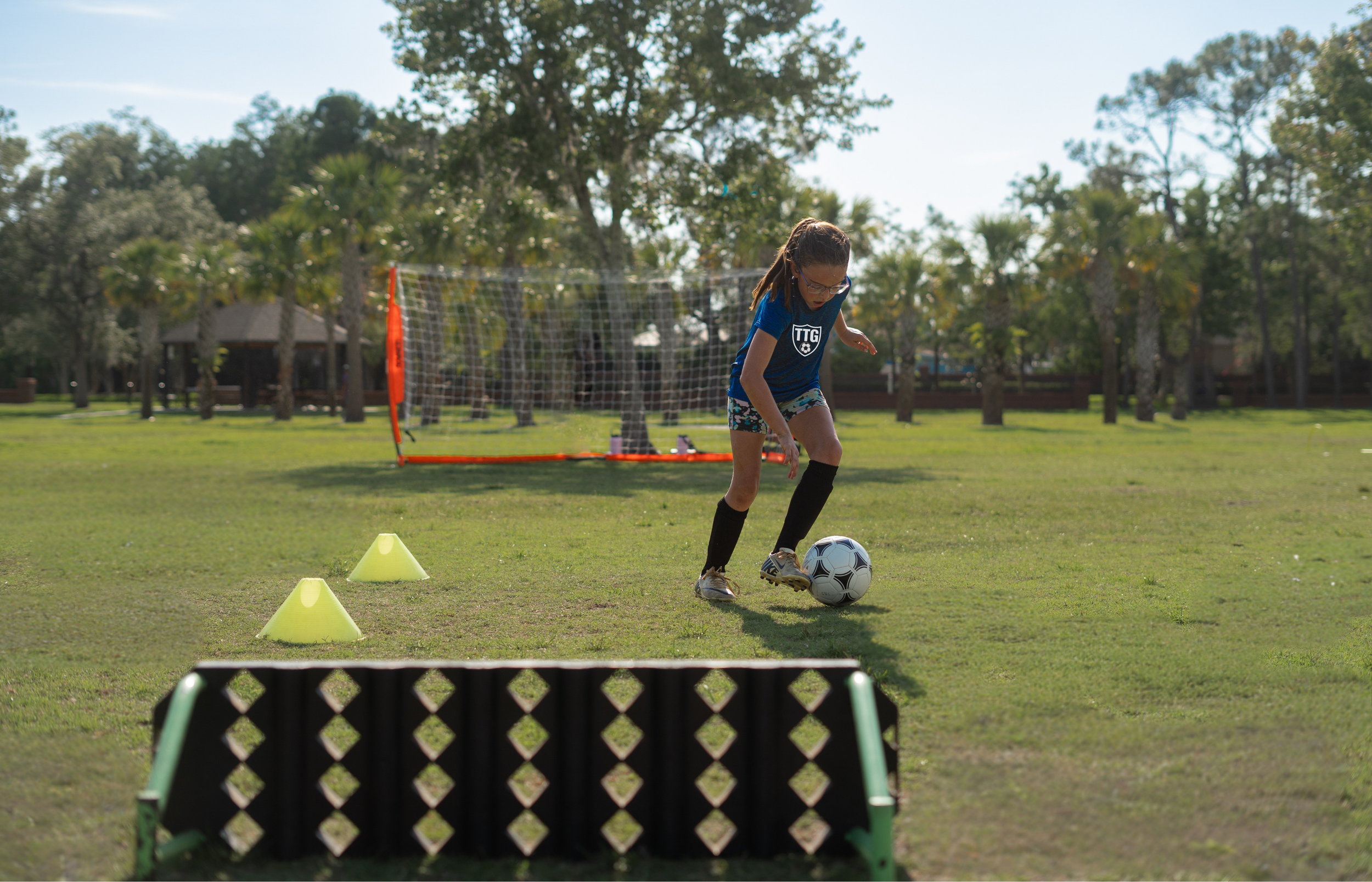 A young girl practicing soccer on a grassy field with orange training cones and a soccer goal in the background. She is wearing a blue sports shirt, black shorts, and black knee-high socks, and is kicking a soccer ball.