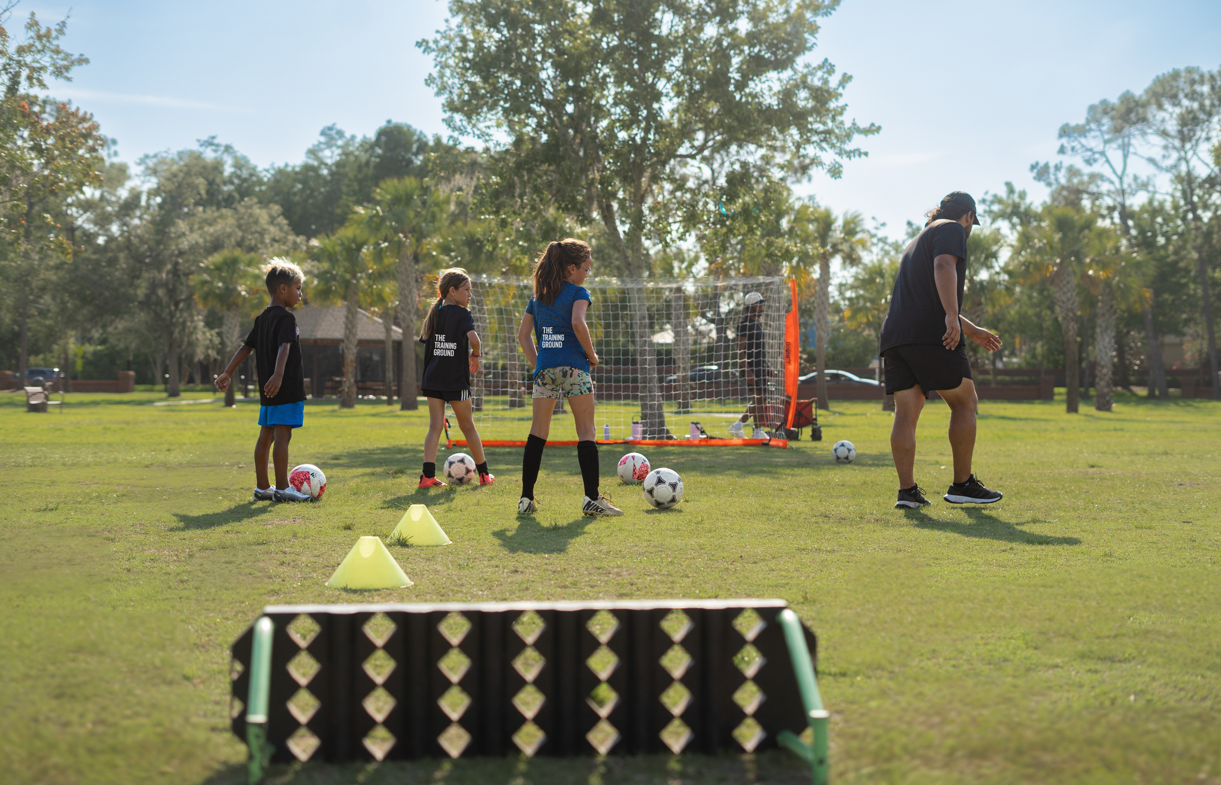 Children participating in a soccer training session outdoors with a coach, standing on grass in front of a small soccer goal, with cones set up for drills.
