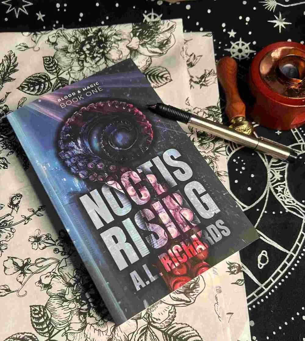 Flat lay of the fantasy romance novel Noctis Rising by A.L. Richarids, featuring a dark celestial book cover with an eye motif, styled on a star-patterned cloth with a pen, wax seal stamp, and floral paper, creating a moody bookish aesthetic.