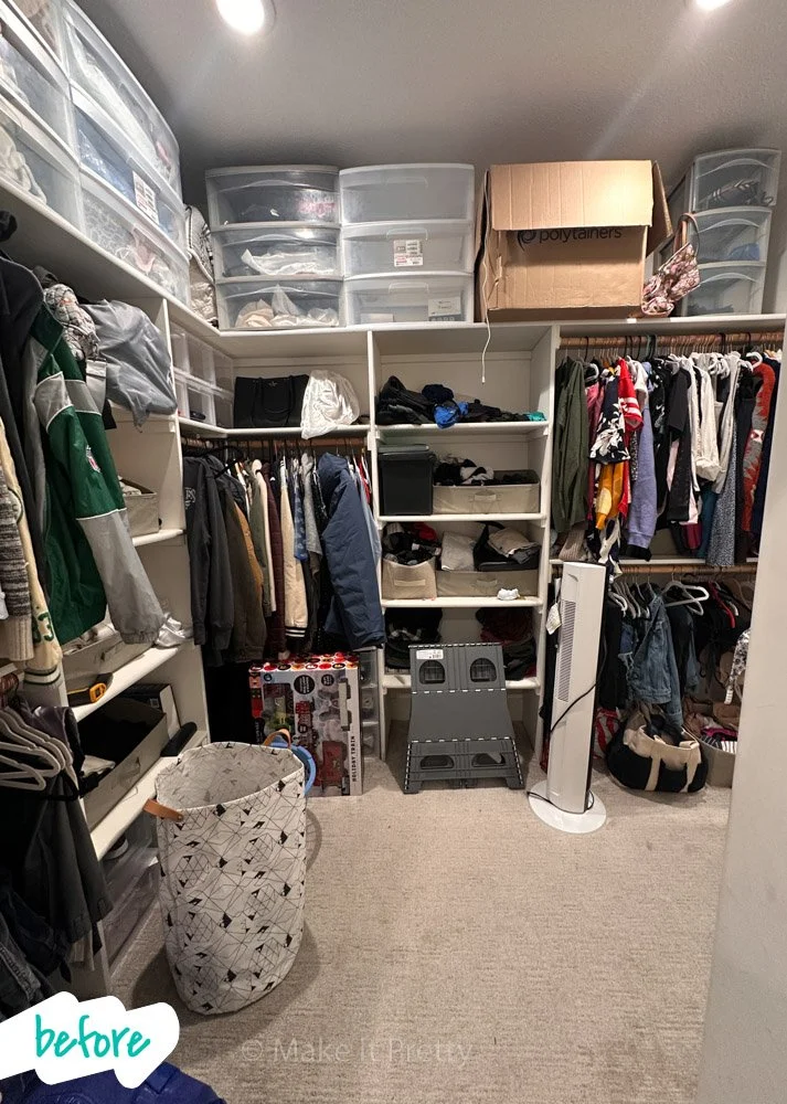 A cluttered walk-in closet with clothes hanging on rods, plastic storage bins on shelves, a cardboard box, and various household items on the floor, including a laundry basket, small step stool, and tower fan.