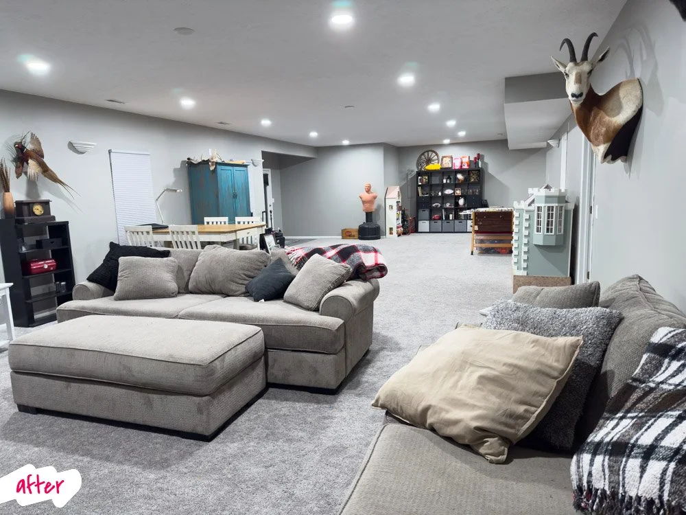 A spacious, well-lit basement living area with beige sofas, an exercise punching bag, a large blue cupboard, a black bookshelf with various items, and a mounted deer head on the wall.