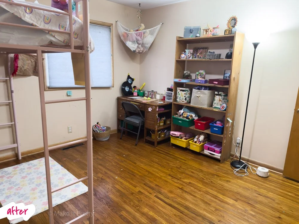A cozy children's bedroom with a pink bunk bed on the left, a small desk near the window, a bookshelf filled with toys and decor, and a wood floor. There is a tall floor lamp on the right and a window with a closed mini blind.