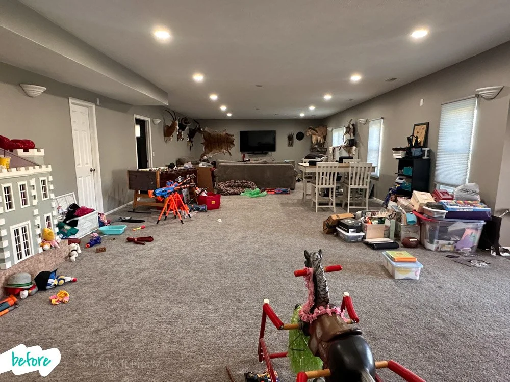 A large basement room filled with children's toys, stuffed animals, and clutter, with a couch, dining table, and mounted animal heads on the walls.