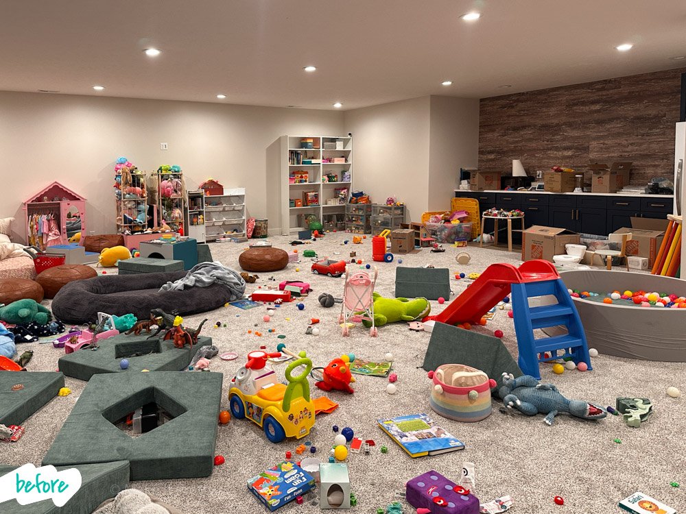 Child's playroom with scattered toys, stuffed animals, a small slide, and disorganized furniture and shelves.