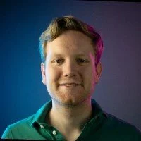 Portrait of a young man with short light brown hair, smiling, wearing a green shirt, against a gradient blue background.