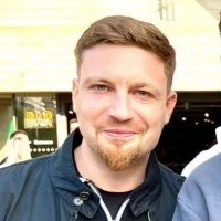 A young man with short brown hair, a beard, and a slight smile, wearing a dark jacket in an outdoor urban setting.
