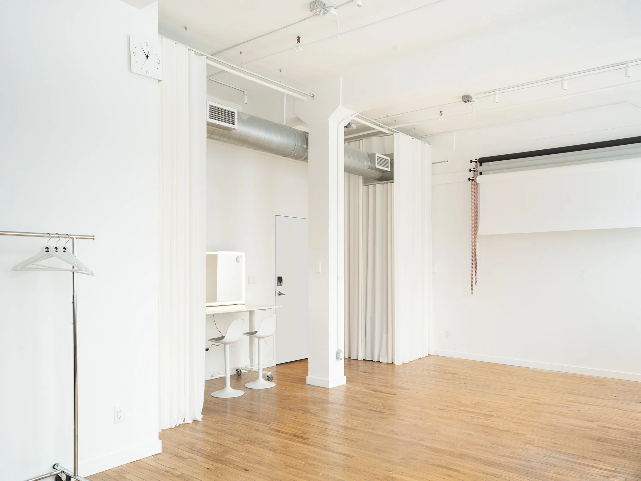 On2, Downtown Toronto's minimalist studio space with wooden floors, white walls, and white drapes. A freestanding clothing rack with hangers and two white swivel chairs at a vanity table are visible.