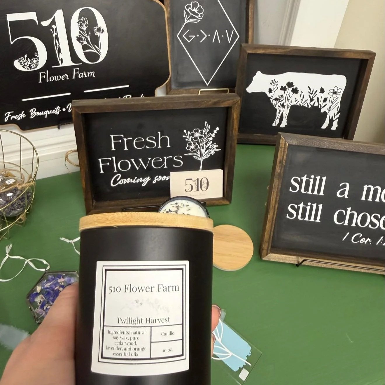 A person holding a candle labeled '510 Flower Farm Twilight Harvest' in front of a display with signs advertising flowers, including one that says 'Fresh Flowers Coming soon' and another with the quote 'Still a moth still chosed' from 1 Corinthians 15. The display includes wooden-framed chalkboard signs with floral and cow designs.