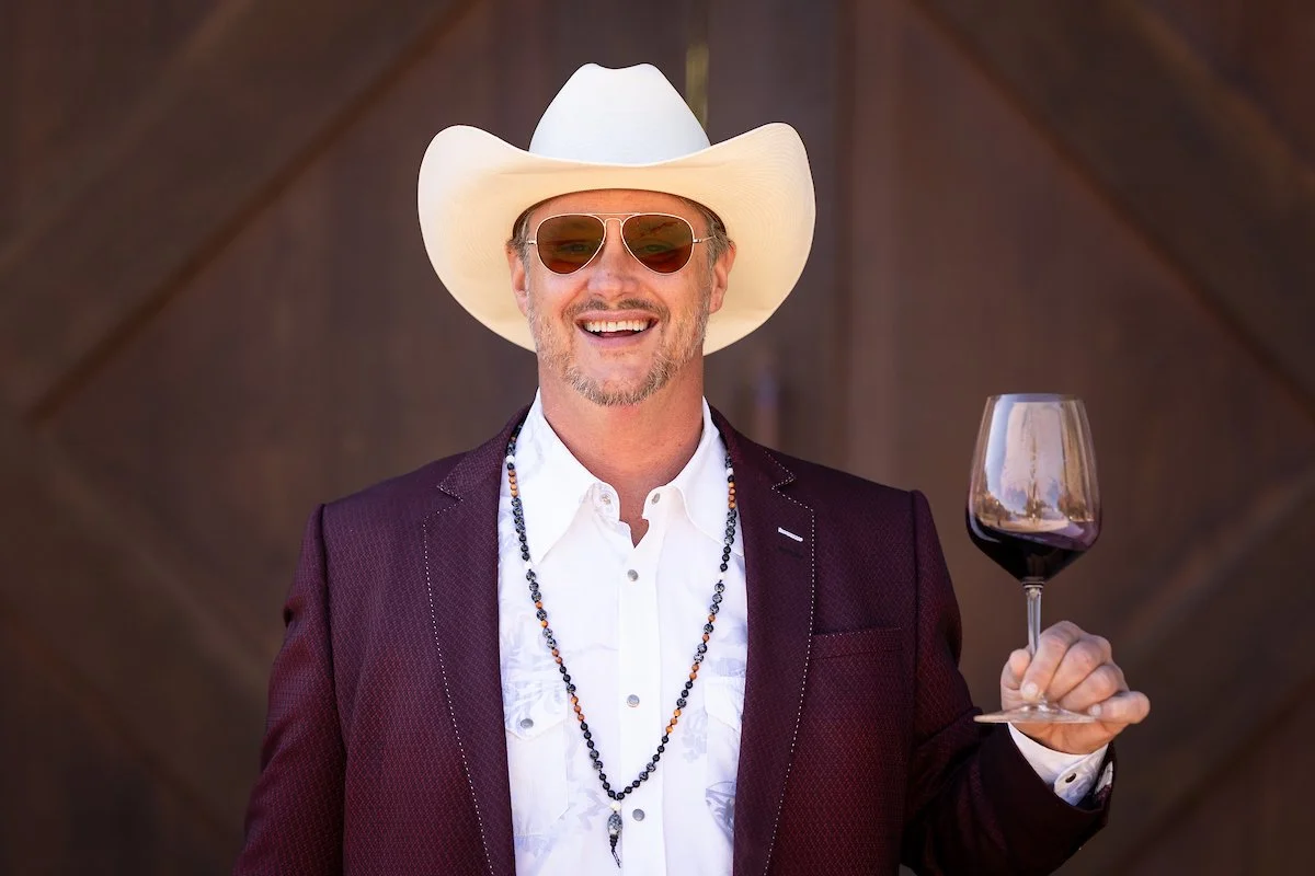 A man with a beard smiling, wearing a white cowboy hat, sunglasses, a burgundy blazer, a white shirt, and beaded necklaces, holding a glass of red wine.