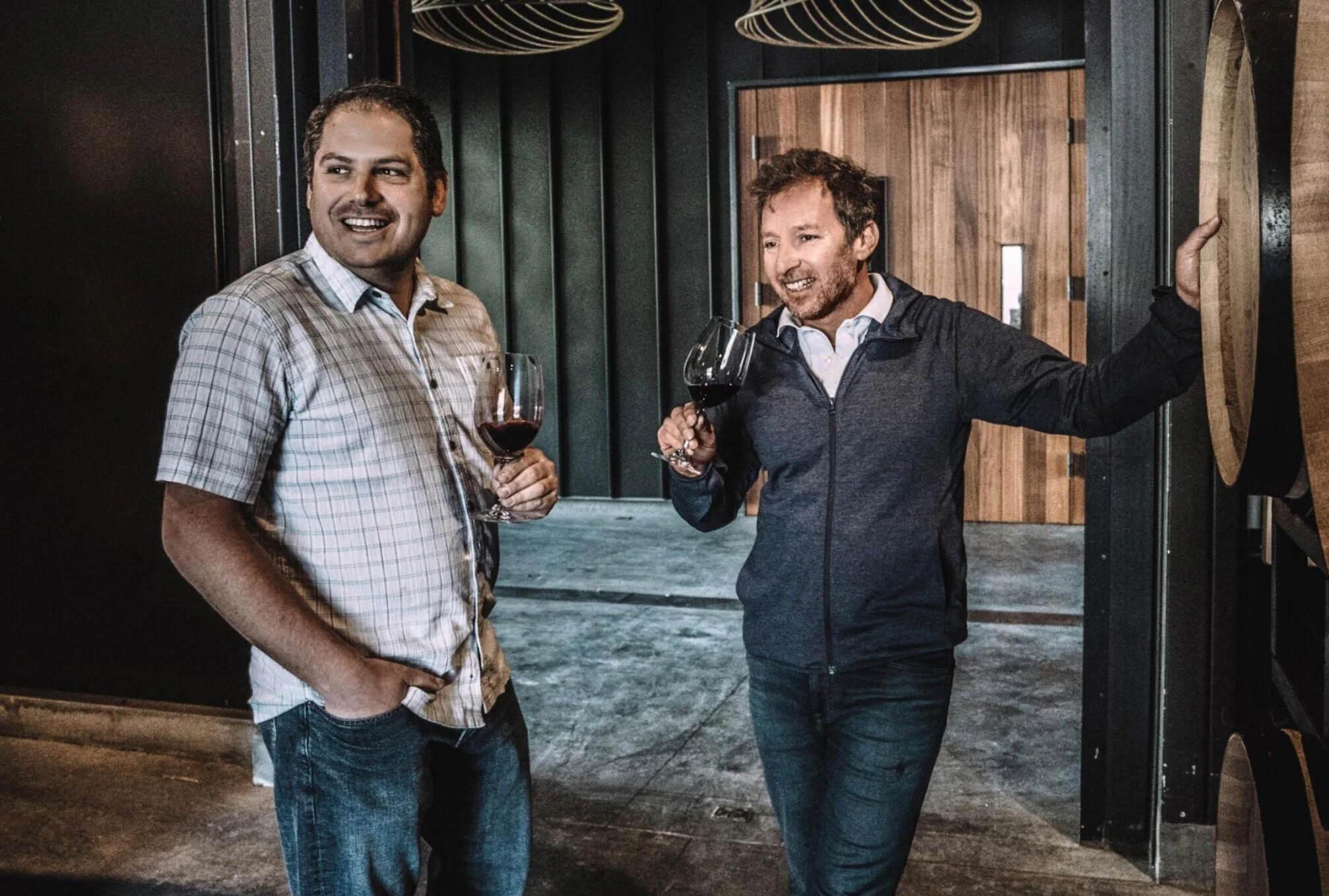 Two men standing near a winery or wine cellar, holding glasses of red wine and smiling, with wine barrels in the background.