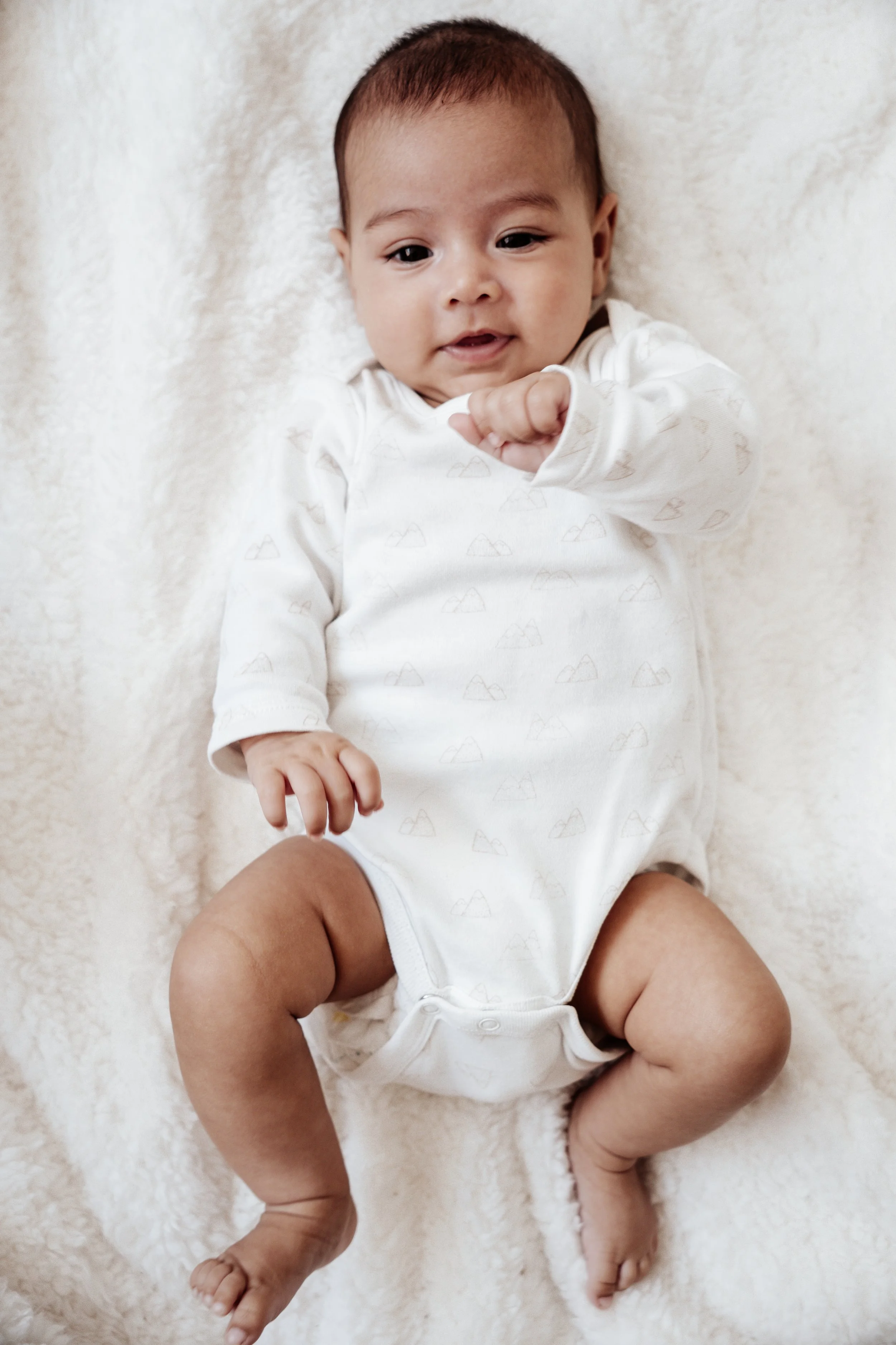 Een baby ligt op een lichtgekleurde, zachte deken en kijkt blij, terwijl het met één hand een vuist maakt en met de andere haar been vasthoudt.