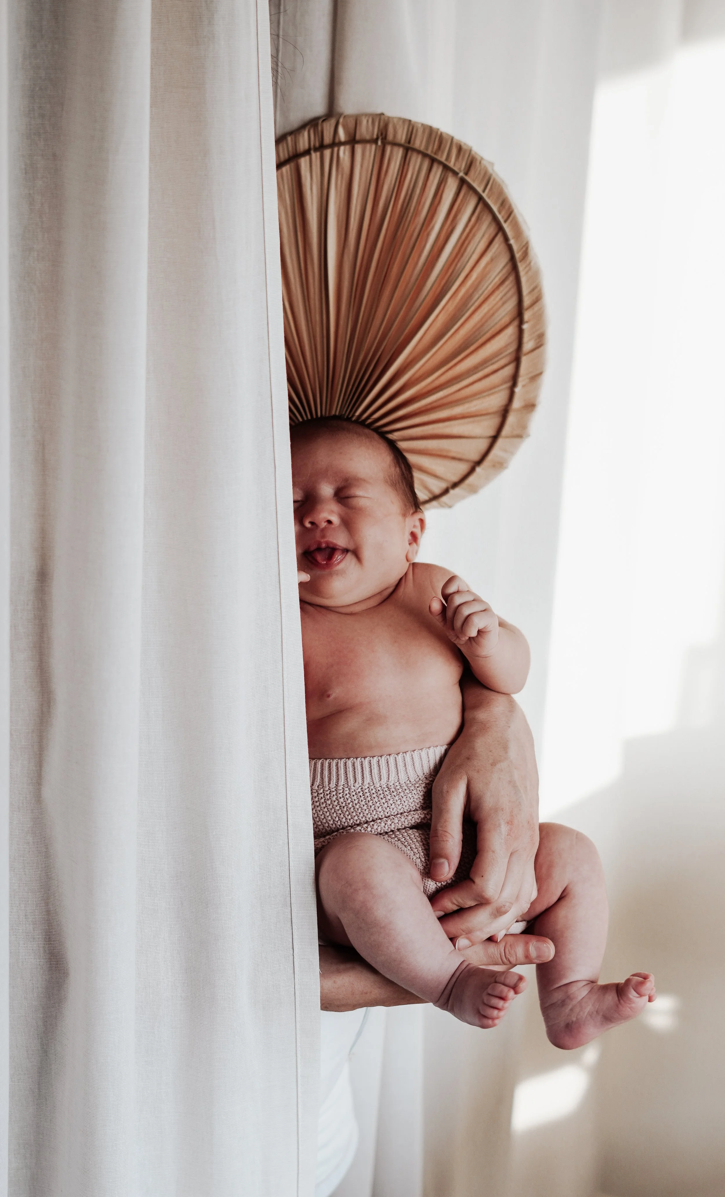 Kijkend naar de foto, een lachende baby die door een persoon wordt vastgehouden, met een rieten parasol achter het hoofd, in een lichte kamer met wit gordijn.