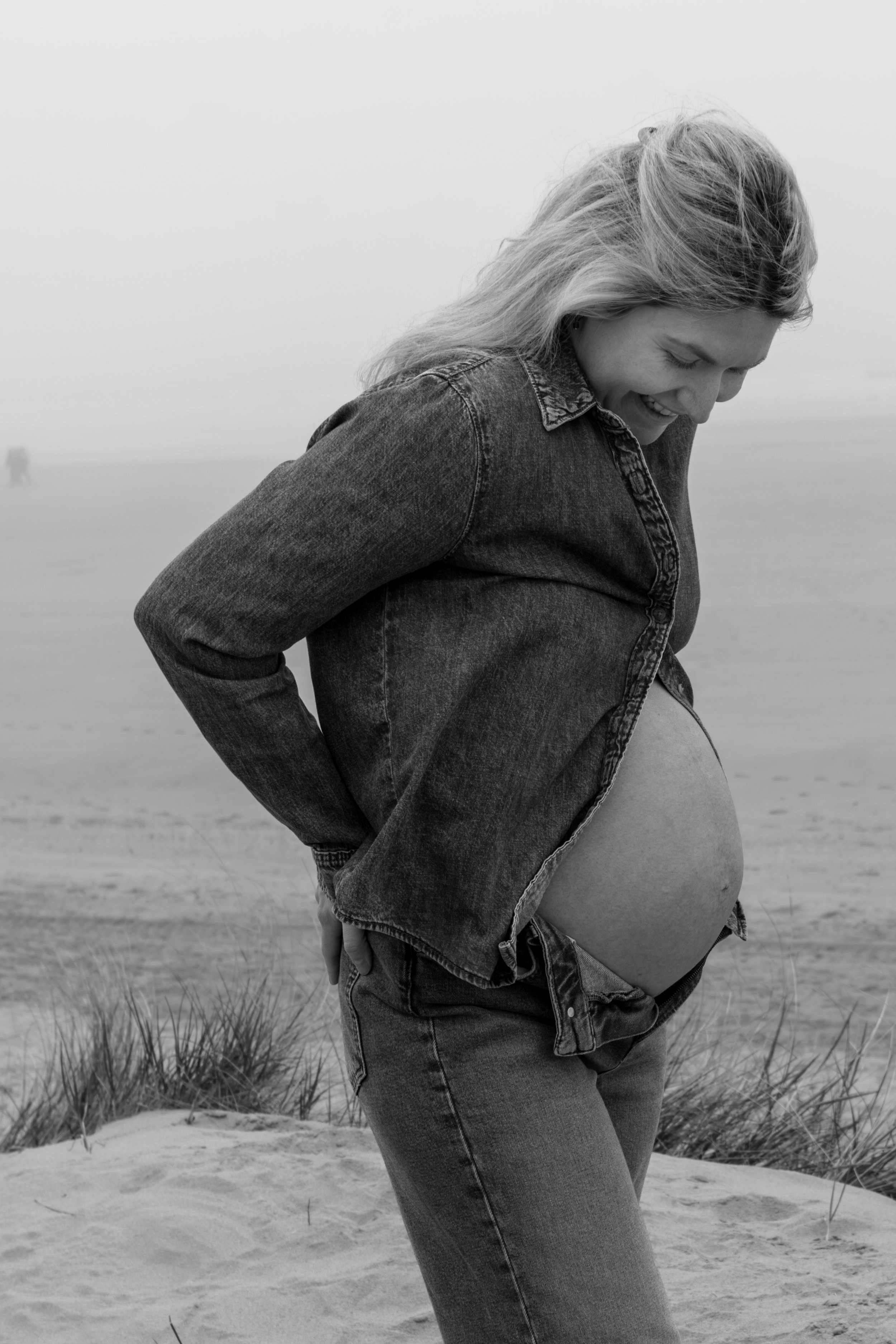 Zwangere vrouw in een denim jas en jeans die haar buik vasthoudt, op een strand met gras en mist op de achtergrond.