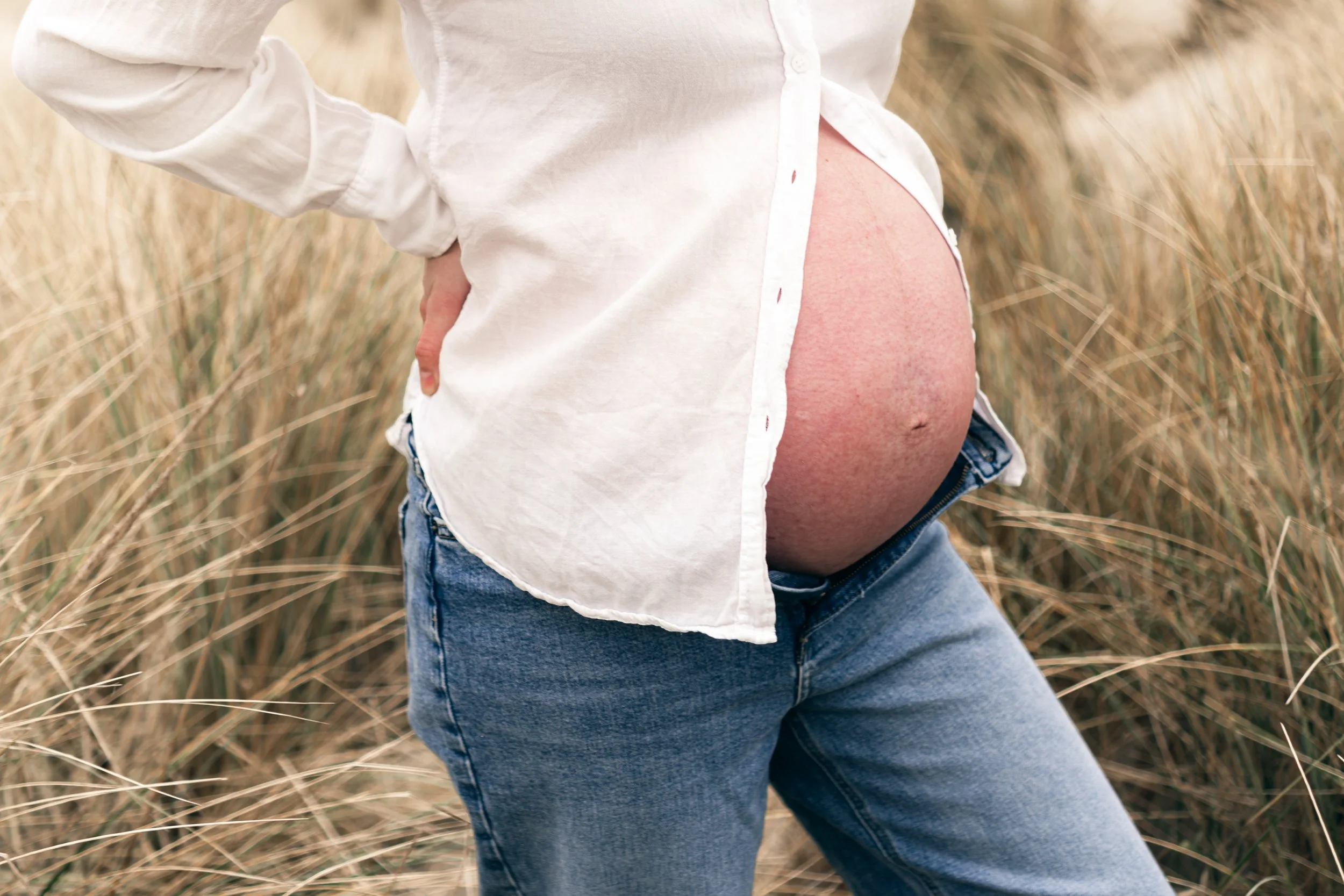 Close-up van een zwangere buik in een witte blouse en blue jeans, in een veld met droog gras.