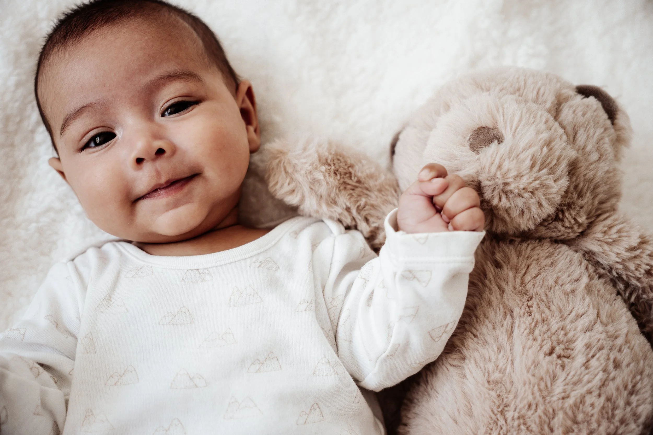 Een baby ligt op een zachte, lichte achtergrond, omringd door een knuffelbeer, glimlachend en naar de camera kijkend.