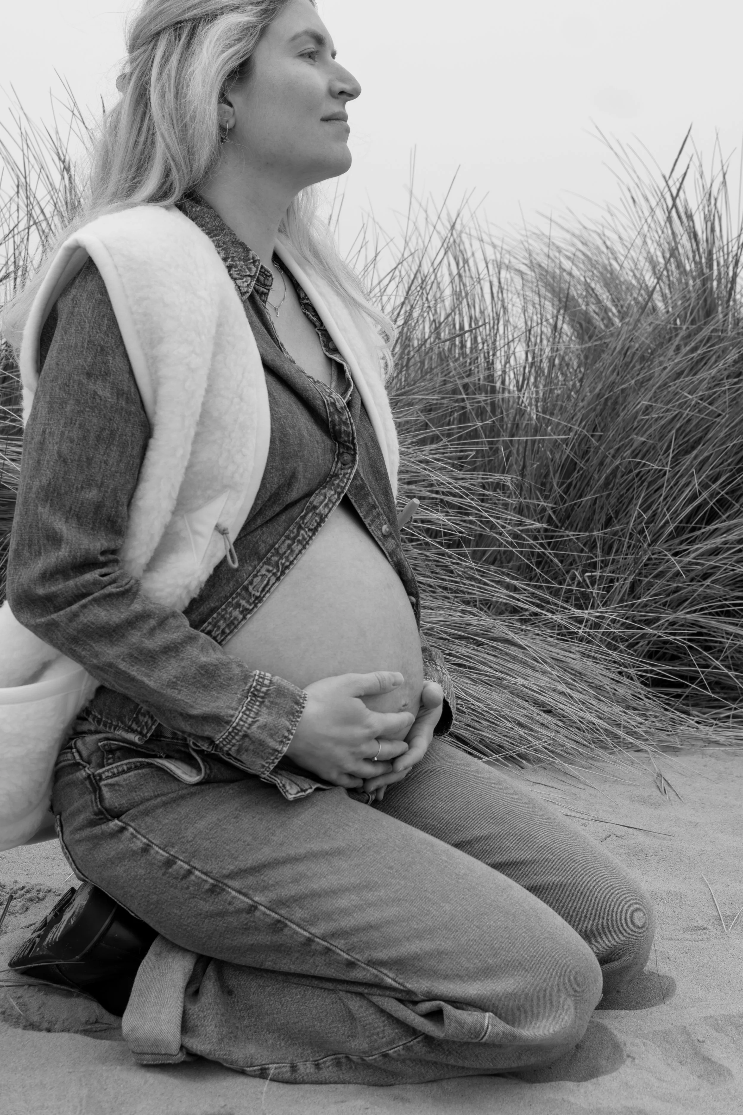 Zittende zwangere vrouw met hand op buik op een zandstrand met gras op achtergrond in zwart-wit.