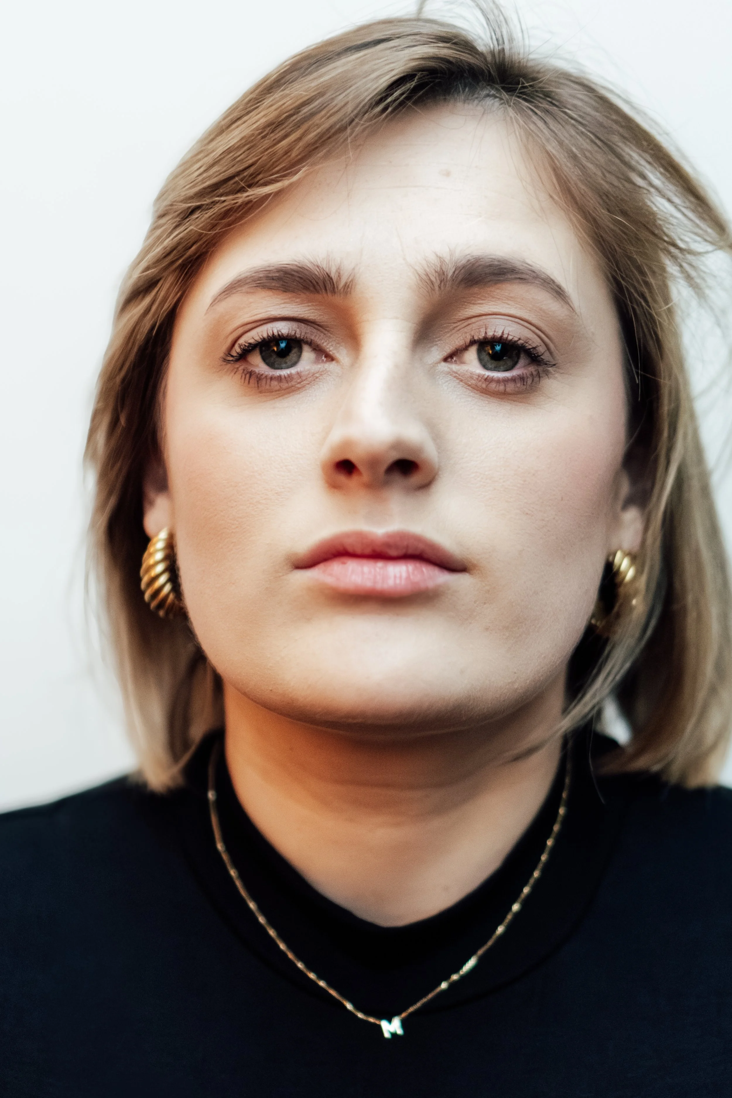 Close-up van een vrouw met bruin haar, lichte huid, draagt gouden oorringen en een ketting met een letter M, tegen een witte achtergrond.
