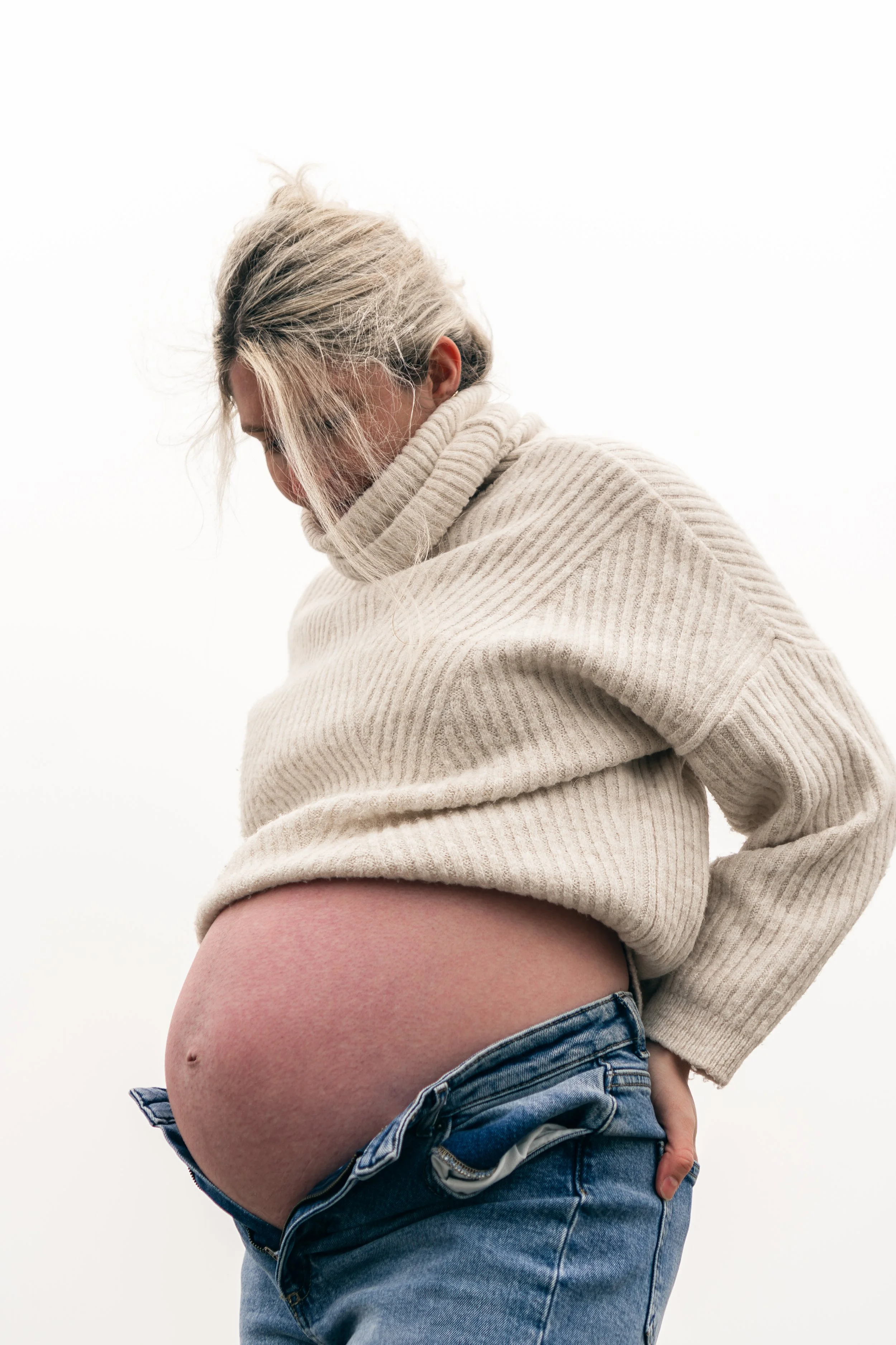 Een zwangere vrouw in een beige trui en denim jeans, die haar buik vasthoudt en naar haar buik kijkt, tegen een witte achtergrond.