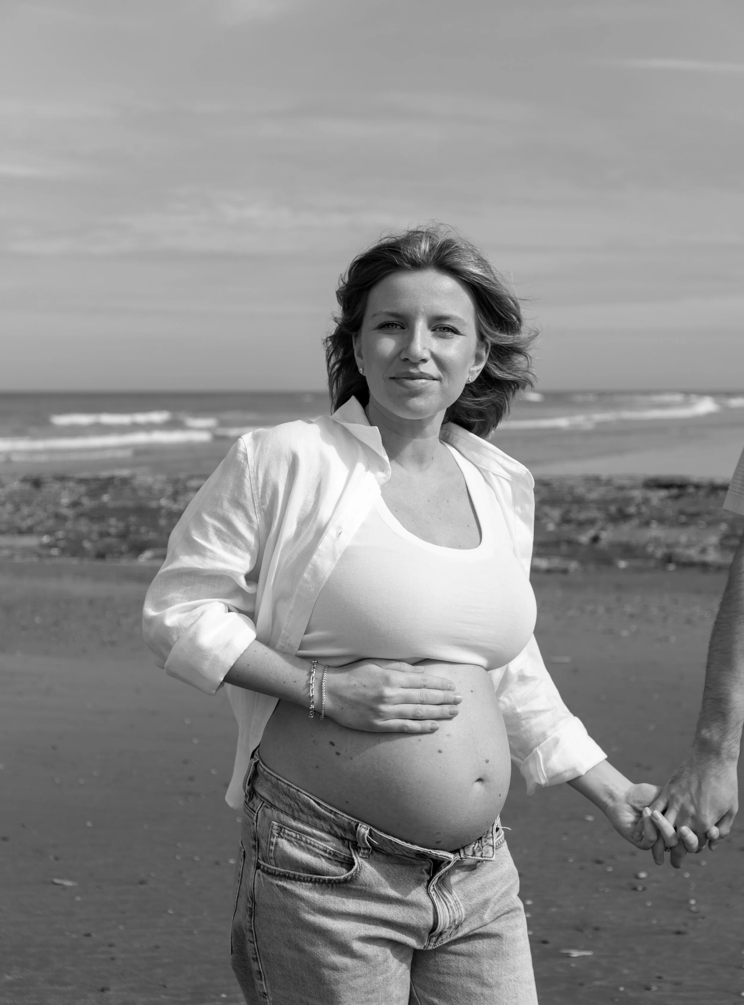 Zwart-wit foto van een zwangere vrouw die op het strand staat, met de oceaan op de achtergrond, lachend en haar buik vasthoudend.