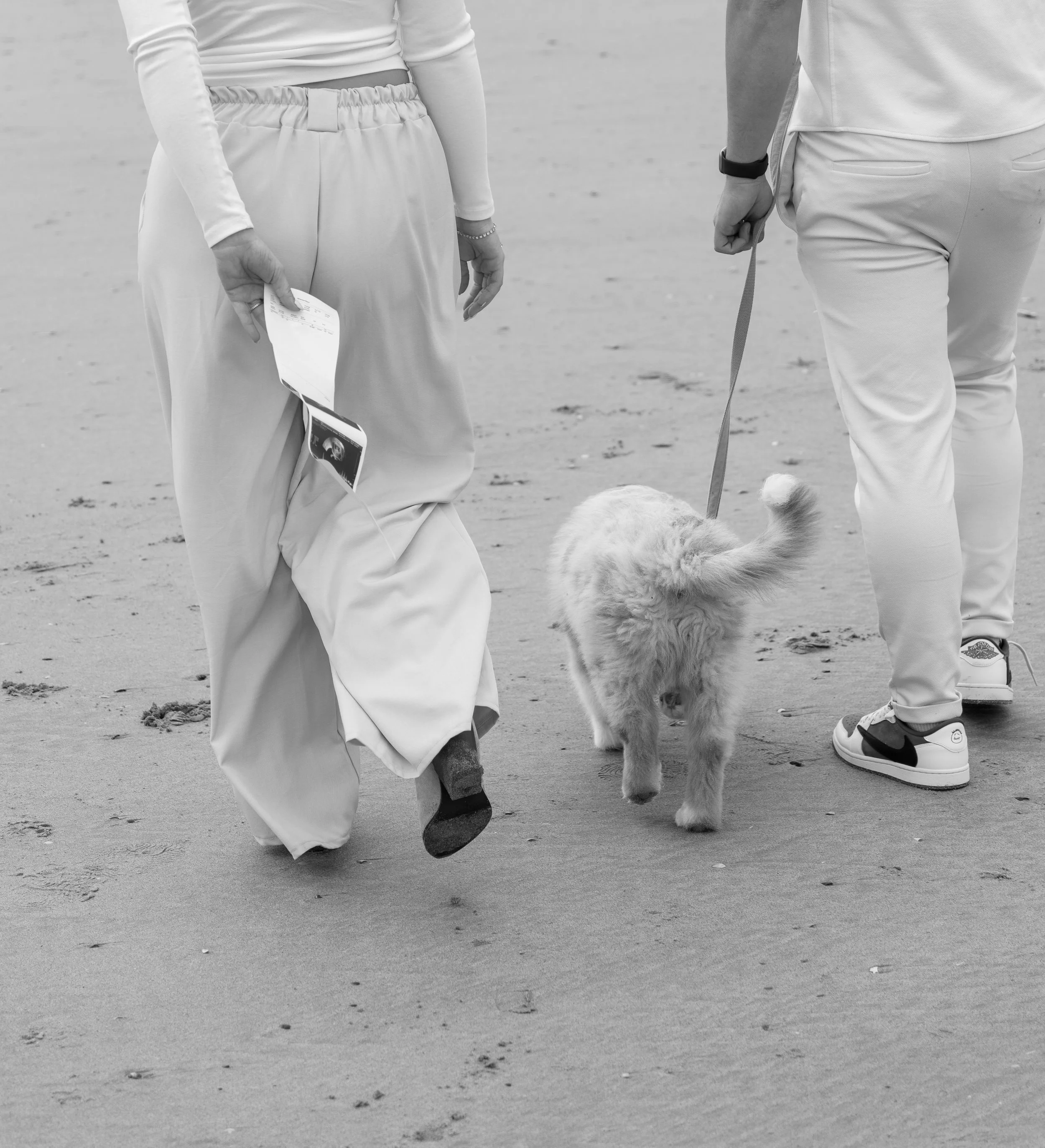 Twee mensen wandelen met een hond op het strand, de foto is zwart-wit.