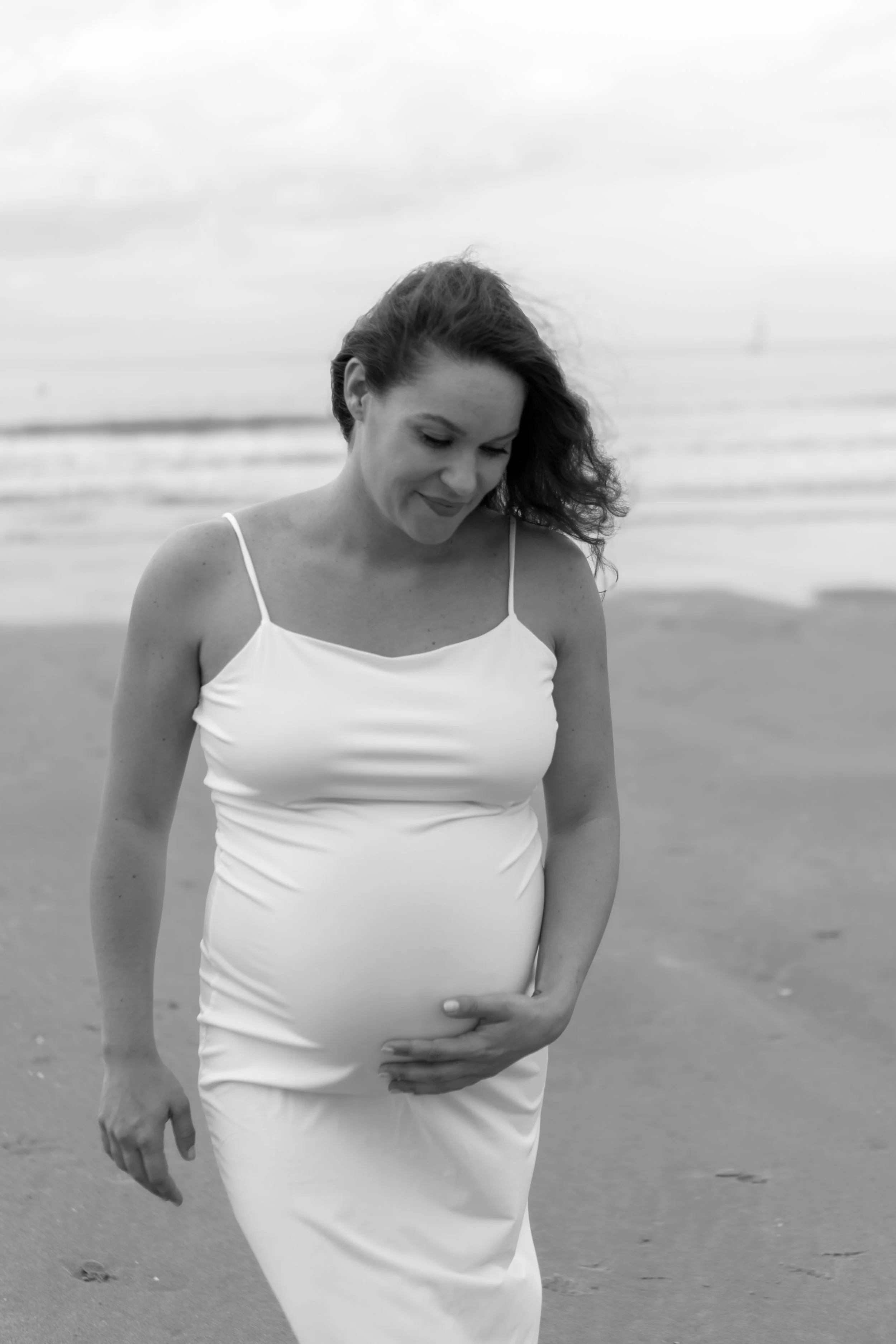 Zwart-wit foto van een zwangere vrouw in een witte jurk, die op het strand staat en haar buik vasthoudt, met de zee op de achtergrond.