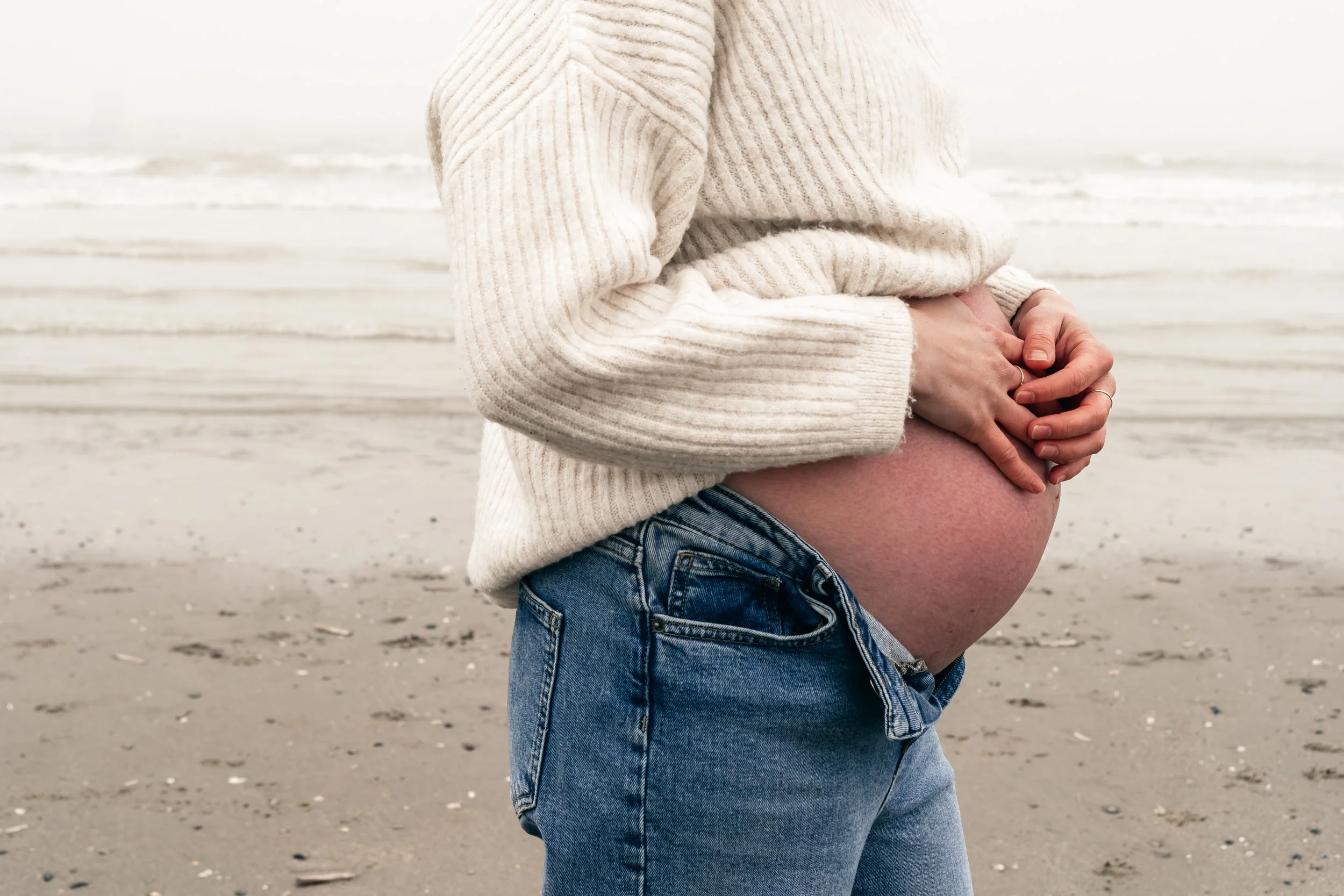 Persoon met een dikke trui die op het strand haar opgezette buik vasthoudt.