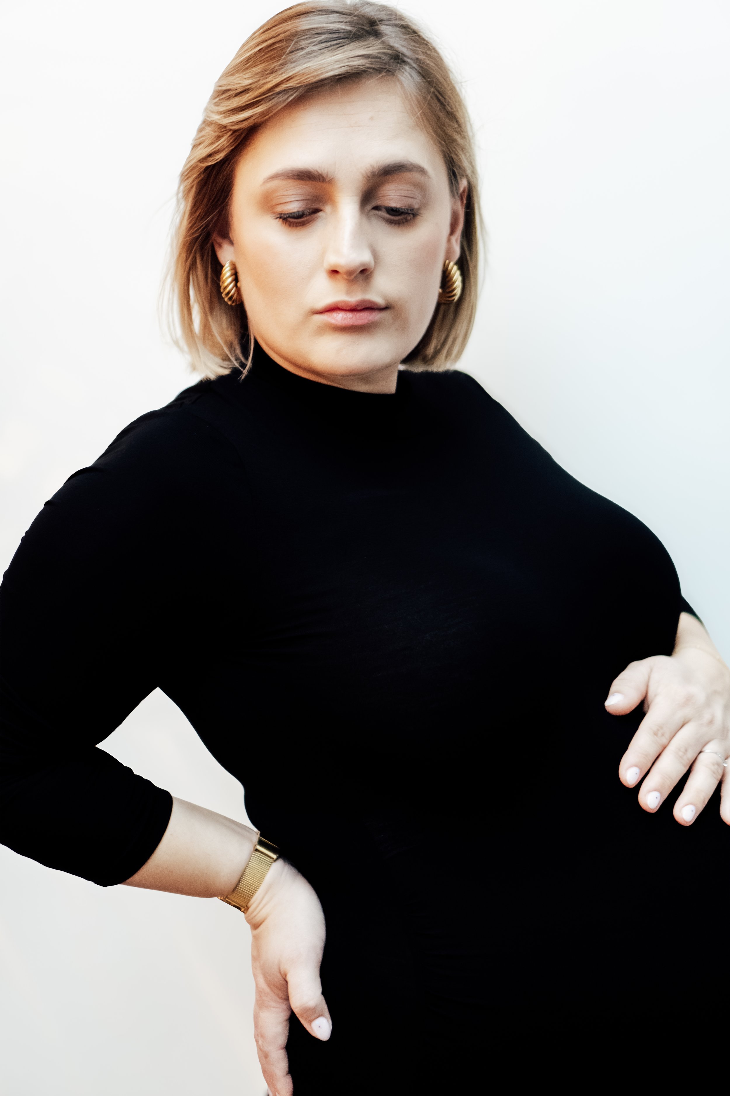 Portret van een vrouw met lichte huid, kort blond haar, goudkleurige oorbellen en een zwarte top, poseert tegen een witte achtergrond.