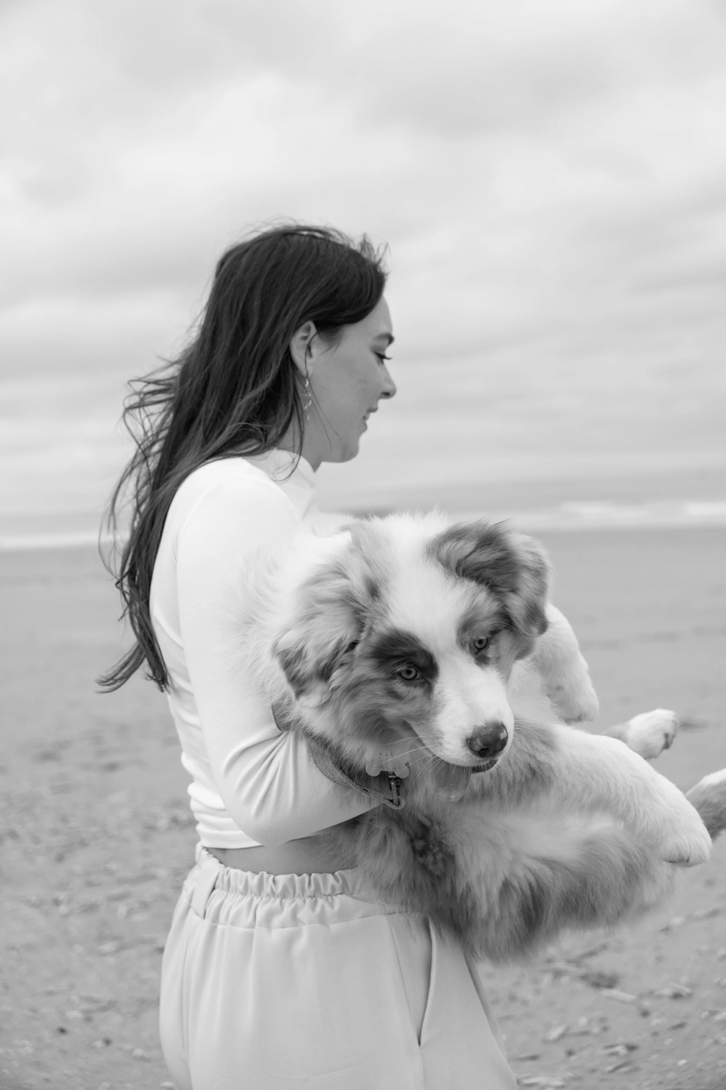 Een vrouw die een grote, witte en grijze Border Collie vasthoudt op een strand met een bewolkte hemel.