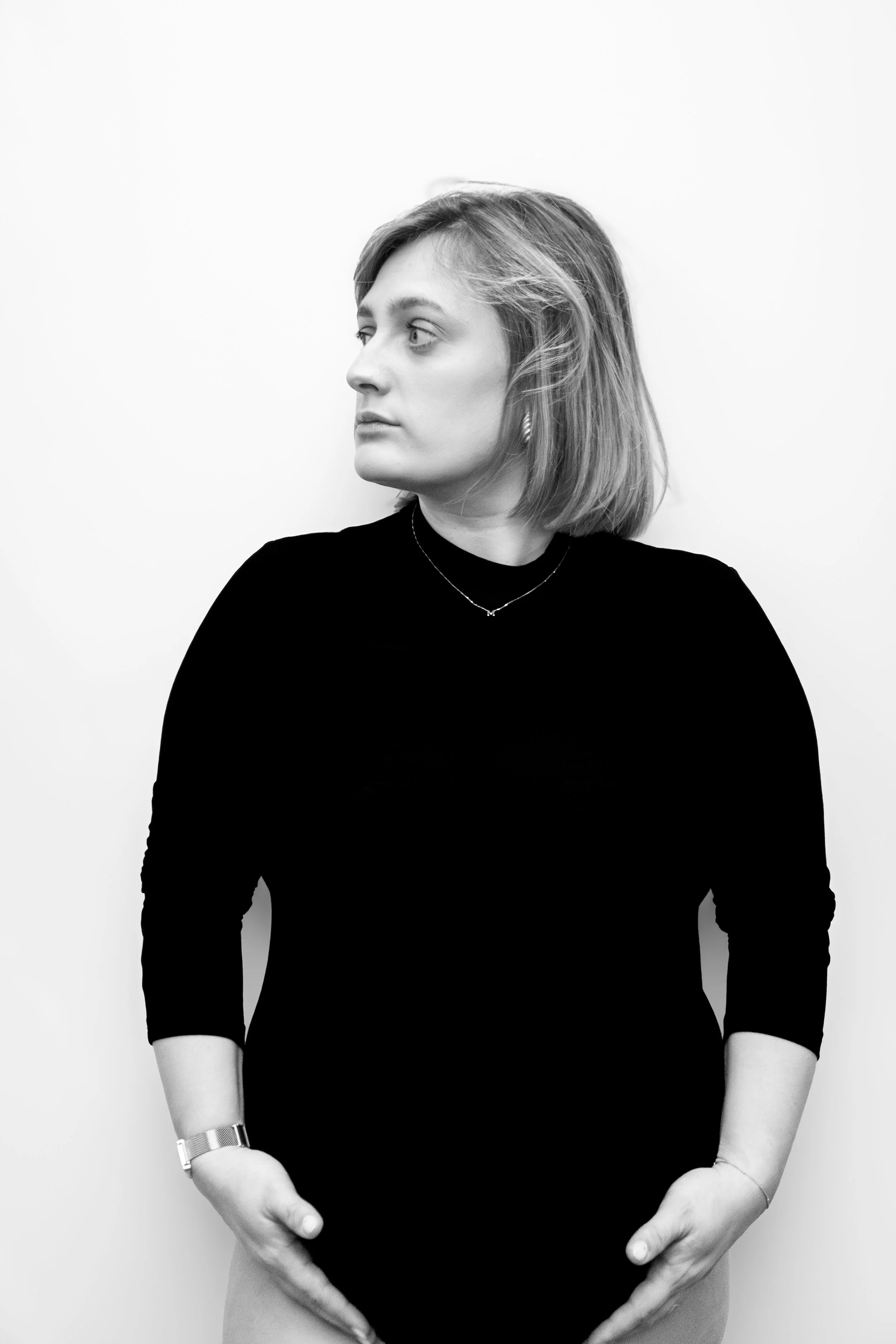 Zwarte en witte foto van een vrouw die naar links kijkt, met haar handen in de vorm van een hart op haar buik, gedragen in een zwarte lange mouwen shirt, met korte haar. 