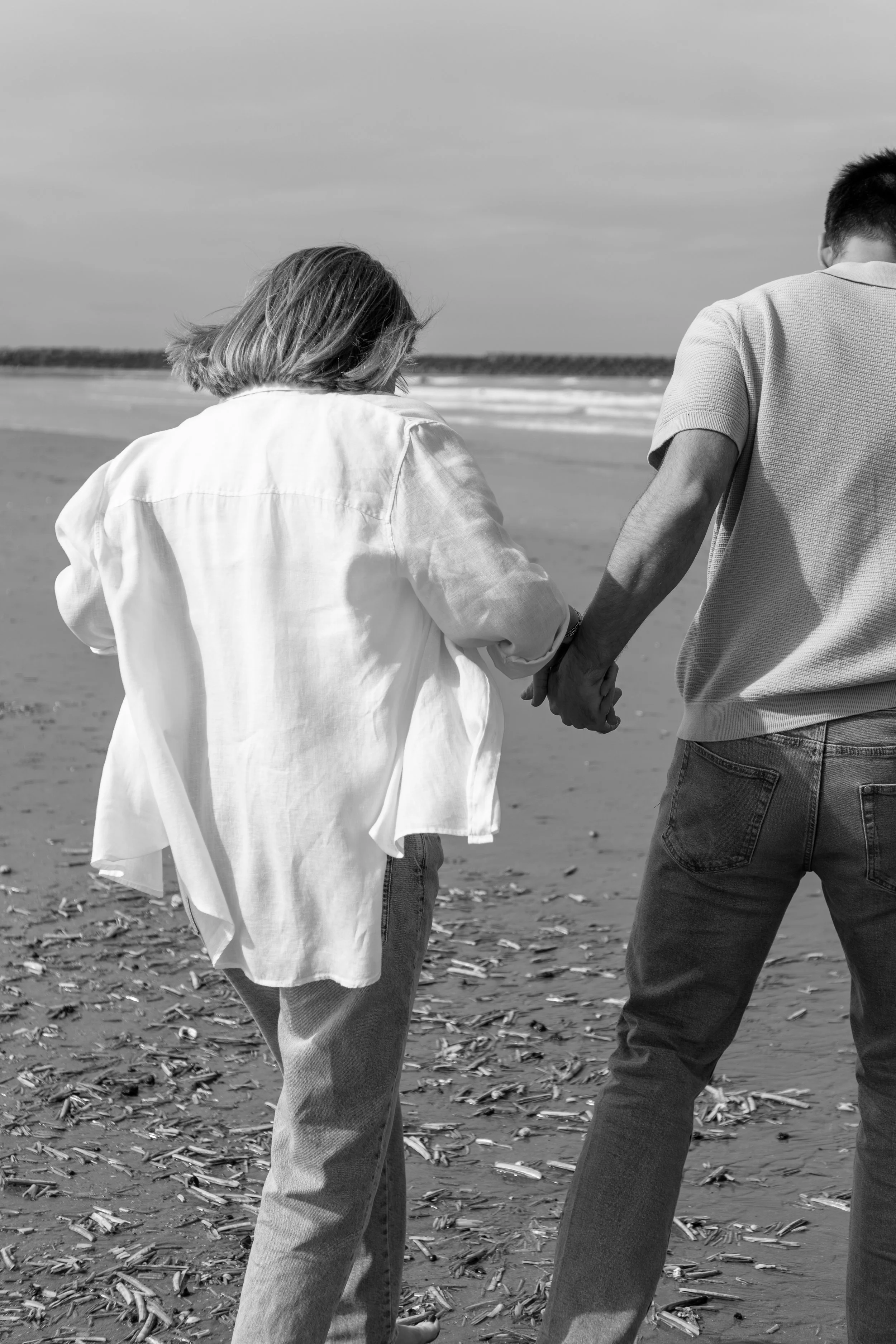 Twee mensen lopen hand in hand op een strand, gezien van achteren, in zwart-wit. Ze dragen casual kleding, één in een witte blouse en de ander in een polo. De zee en de lucht vormen de achtergrond.