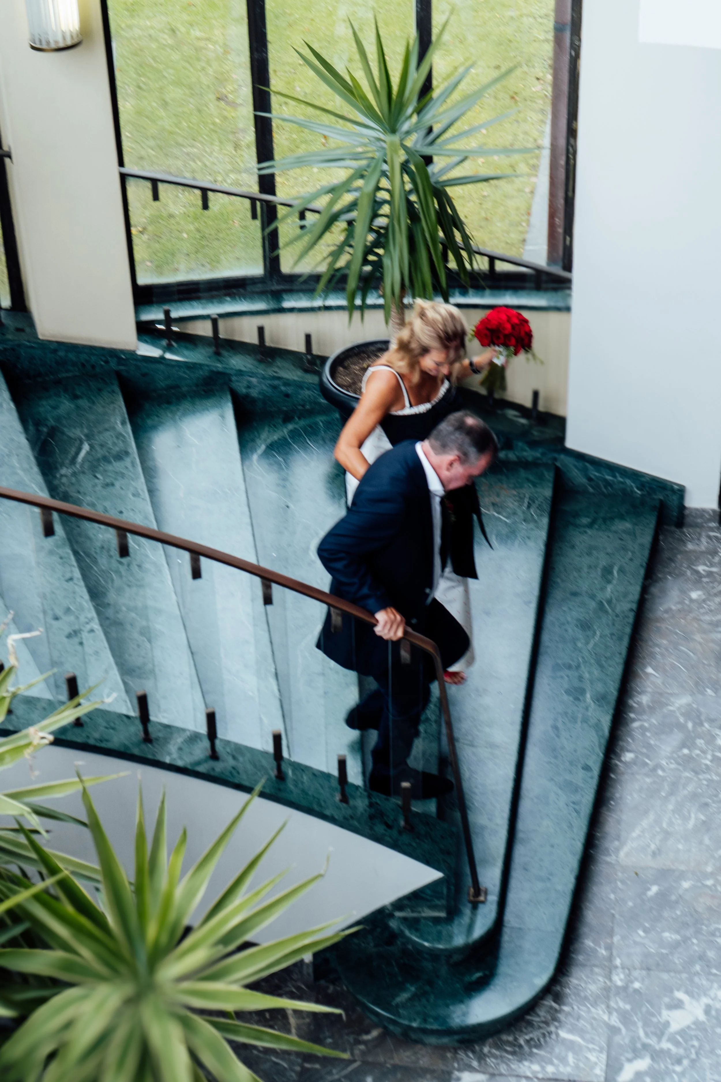 Een vrouw en een man lopen samen de trap op in een gebouw met grote ramen en veel groen buiten. De vrouw houdt een rode bos bloemen vast.