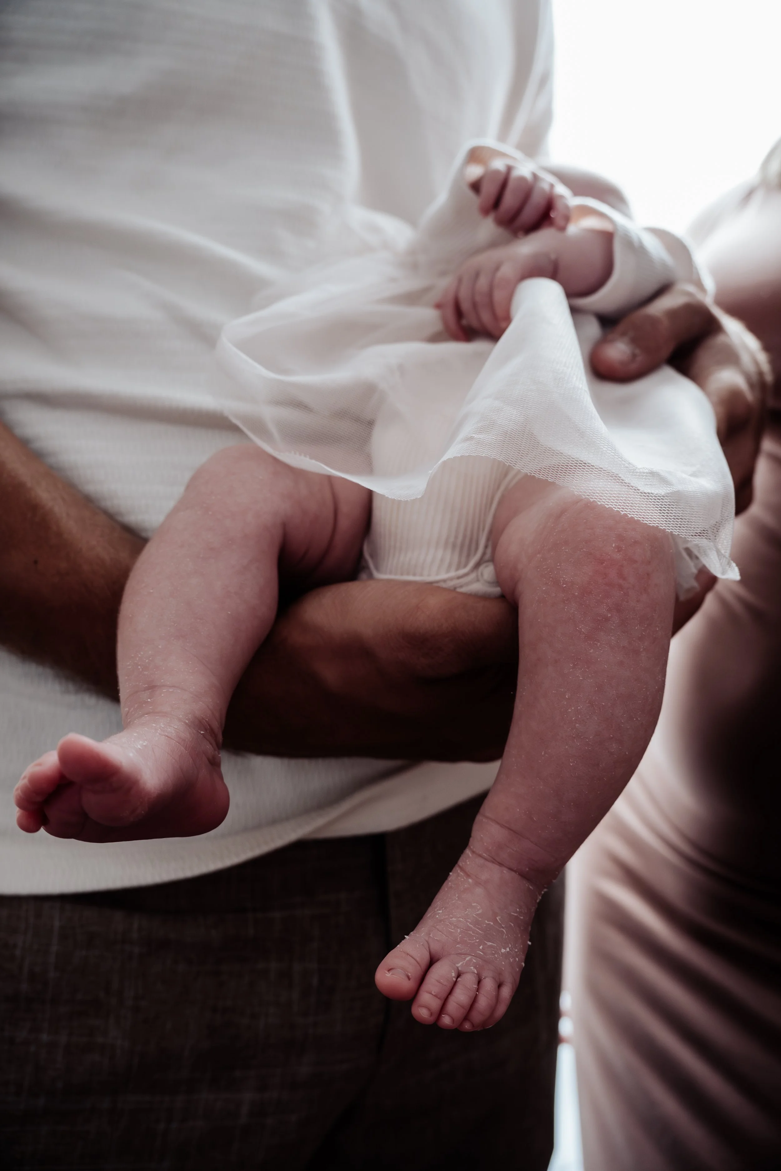 Close-up van een baby die wordt vastgehouden door een volwassene, de baby ligt op de buik en draagt een wit luier en een wit shirtje.