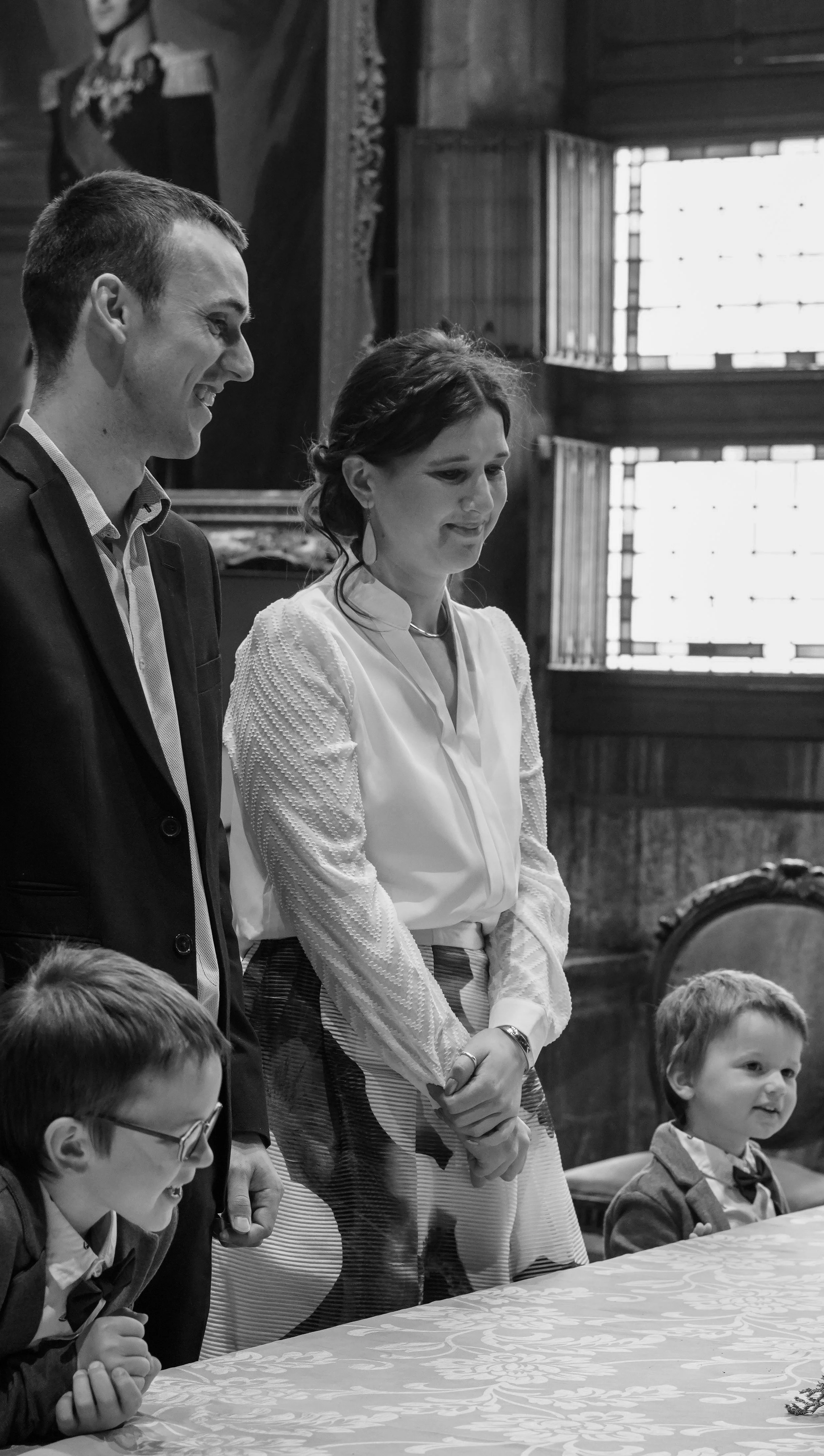 Zwart-wit foto van een groep mensen, inclusief vier jonge jongens en twee volwassenen, die bij een tafel staan in een houten zaal. De volwassenen lijken een gesprekje te voeren. Op de achtergrond een raam en een kerkelijk kunstwerk.