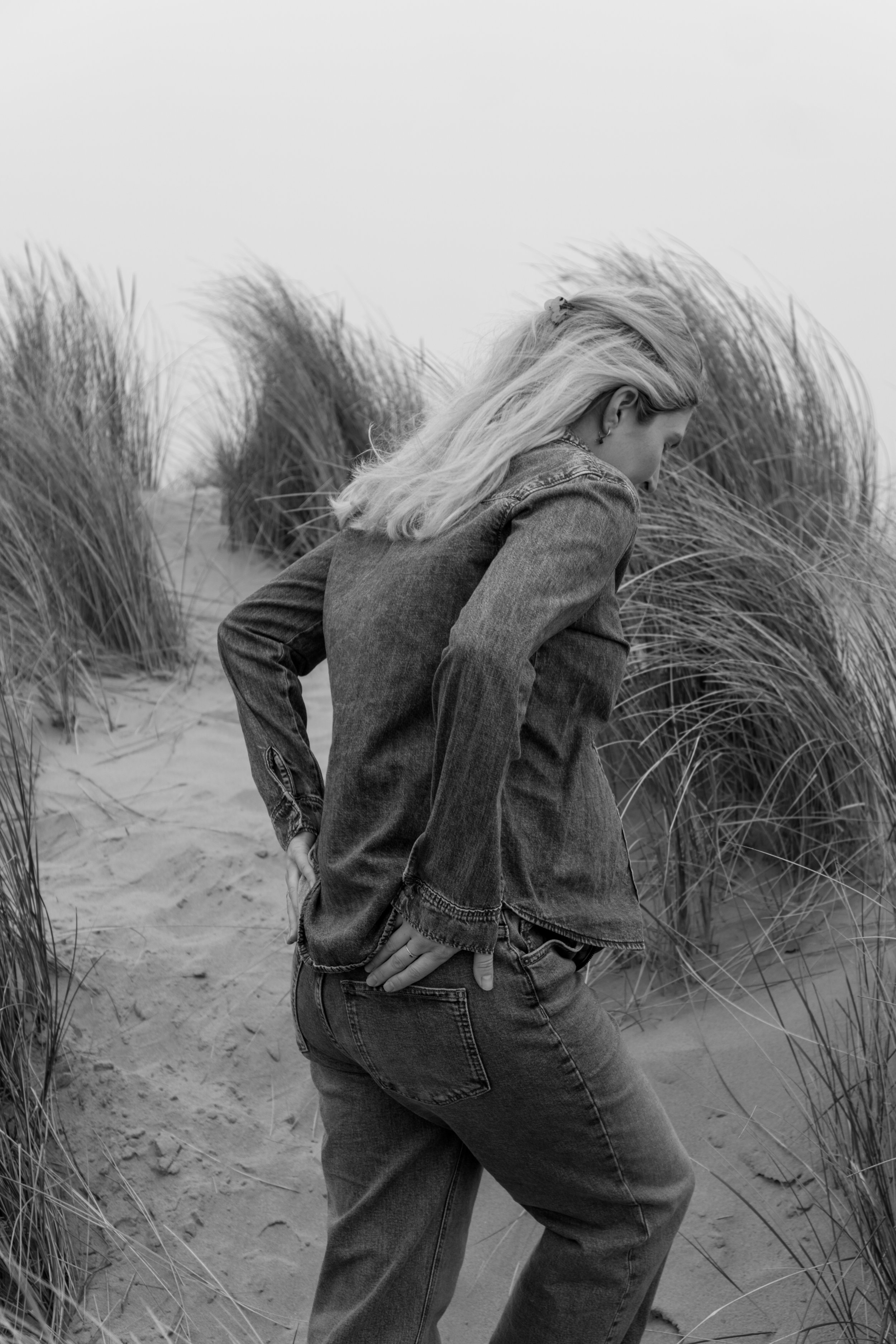 Een vrouw met blond haar, gekleed in een denim jas en jeans, staat op een zandstrand tussen grasachtige planten, met haar hoofd naar beneden en haar handen op haar heupen.