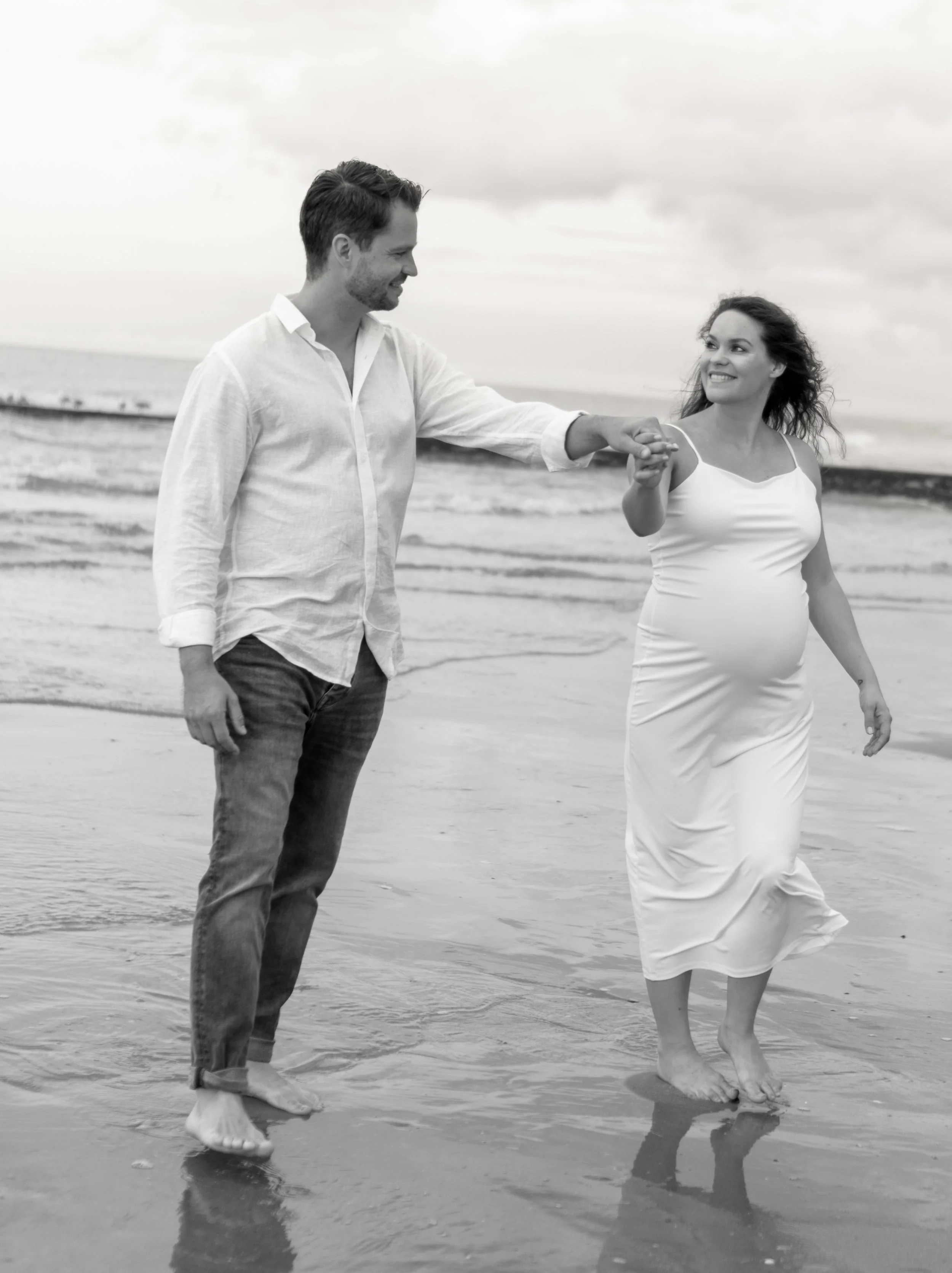 Een man en een vrouw lopen hand in hand op het strand, de man draagt een witte overhemd en de vrouw een witte jurk, beiden zijn met blote voeten in het water.